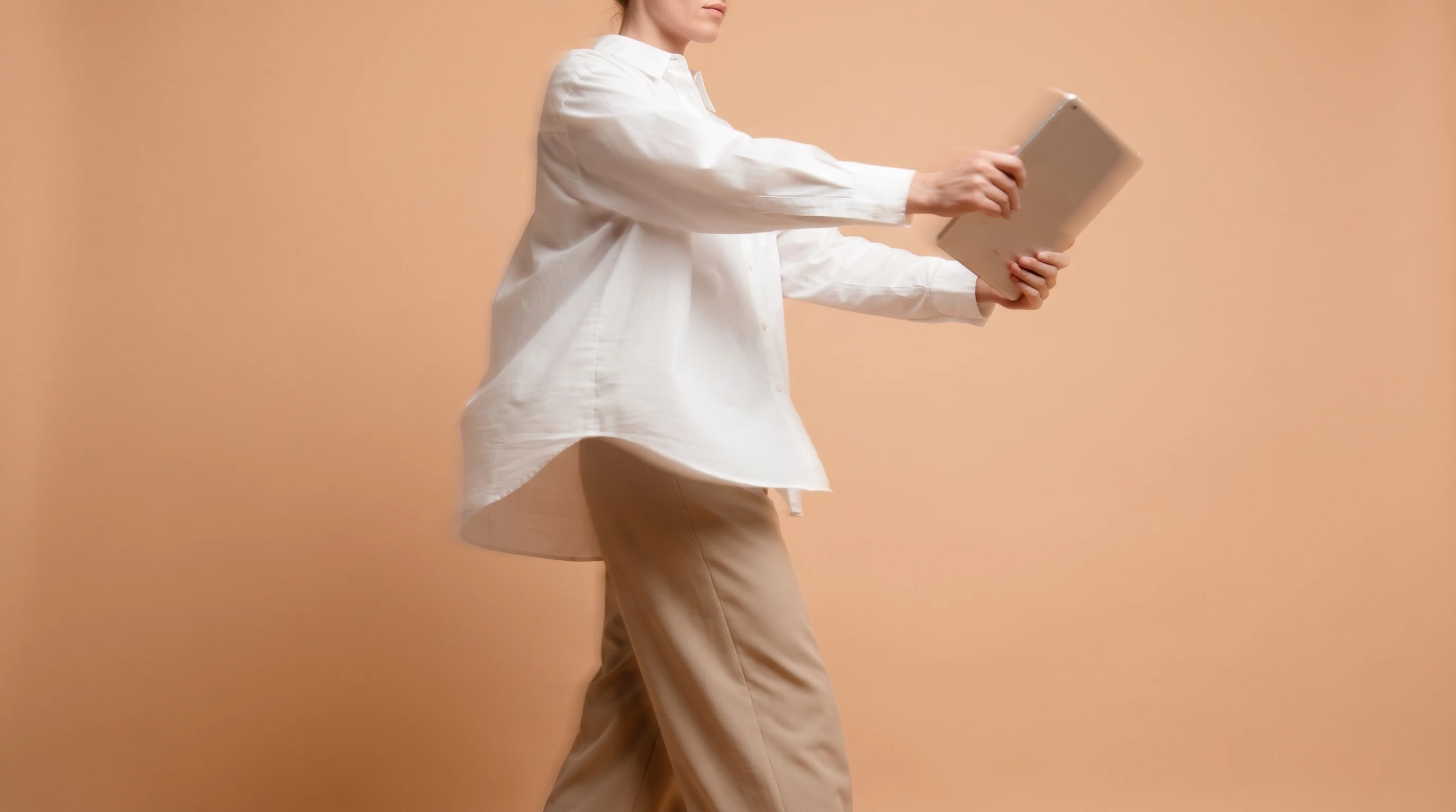 a person mid-spin in a peach studio backdrop, arms extended holding an unbranded tablet angled toward camera, outfit a crisp white oversized shirt