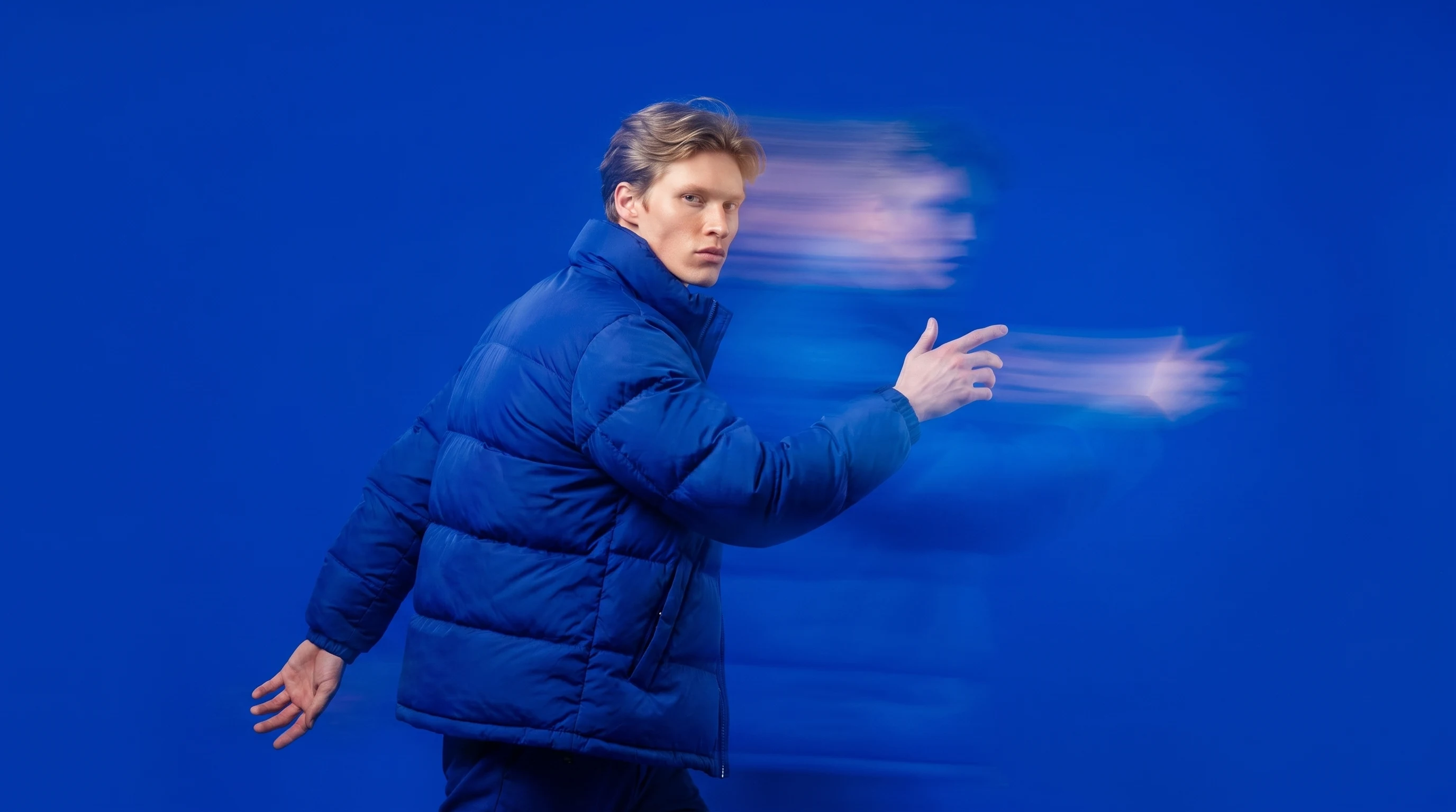 A person mid-step through a vivid cobalt-blue studio, one hand gesturing at an invisible screen beside them, torso turned three-quarter toward camera