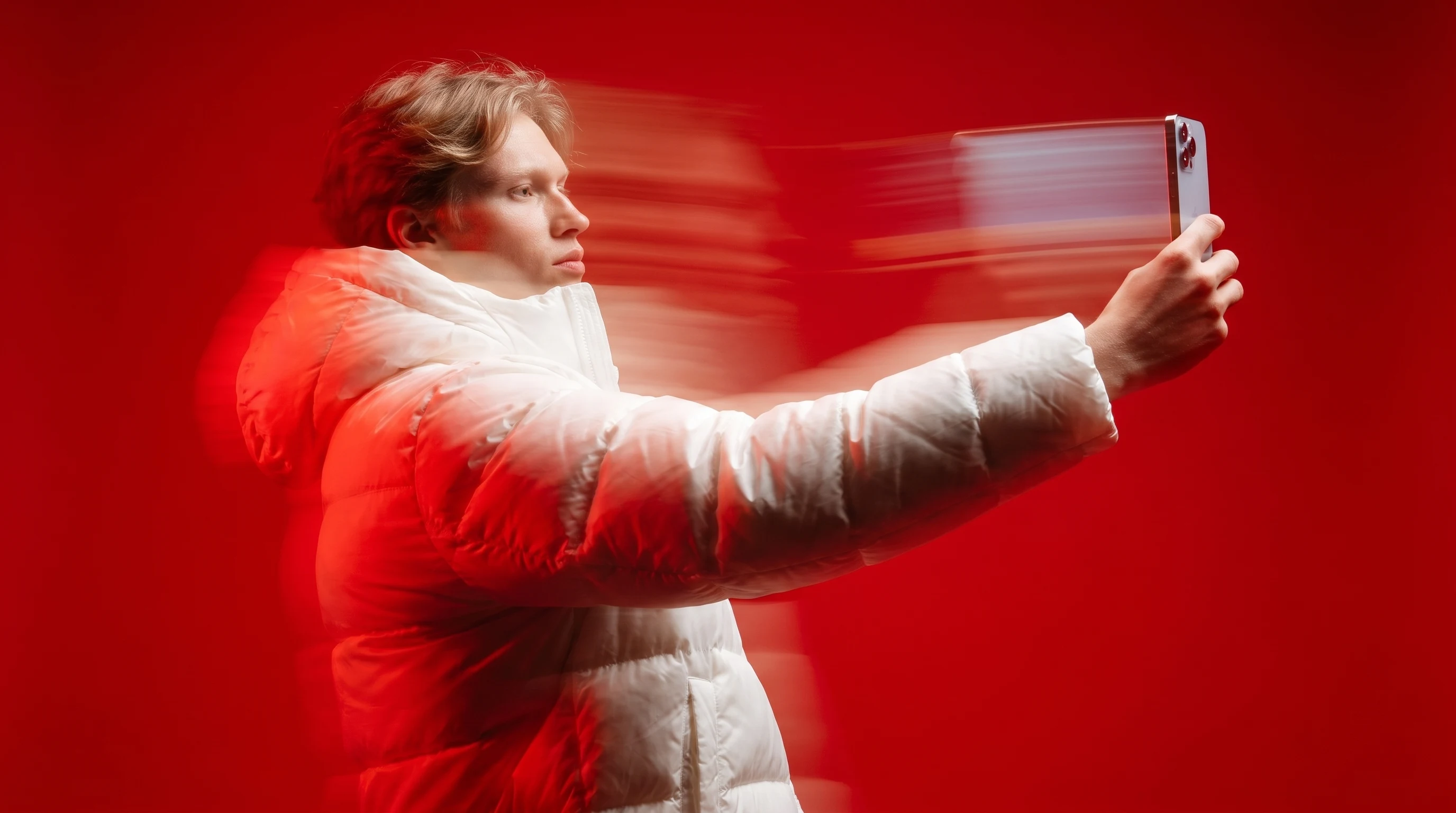 A person mid-spin holding a smartphone toward camera in a bold saturated red studio, arm extended, capturing a vertical selfie-style frame
