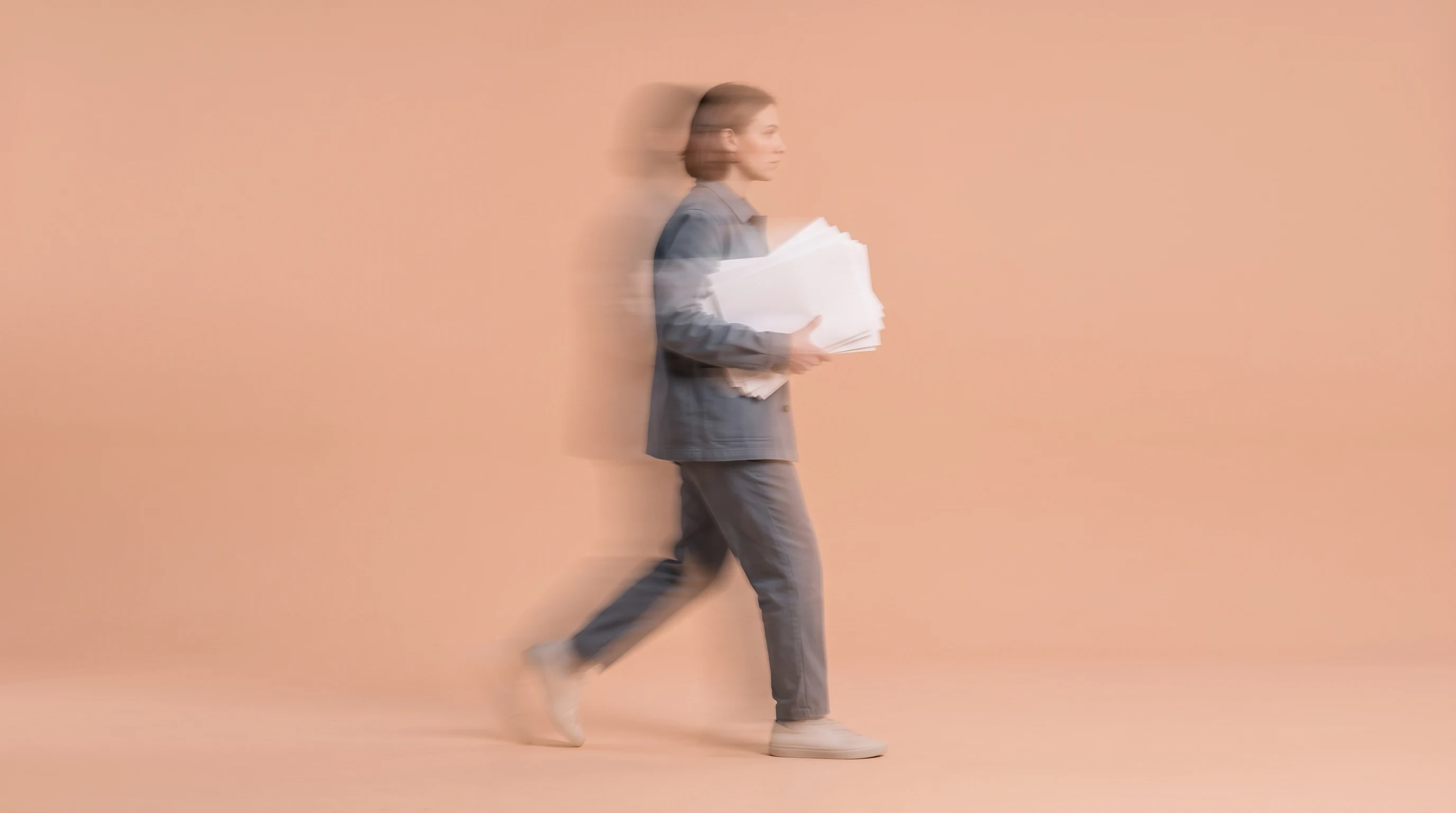 Person mid-step across a peach-backdrop studio space, profile framing, cradling a stack of unbranded printed concept sheets against their chest,