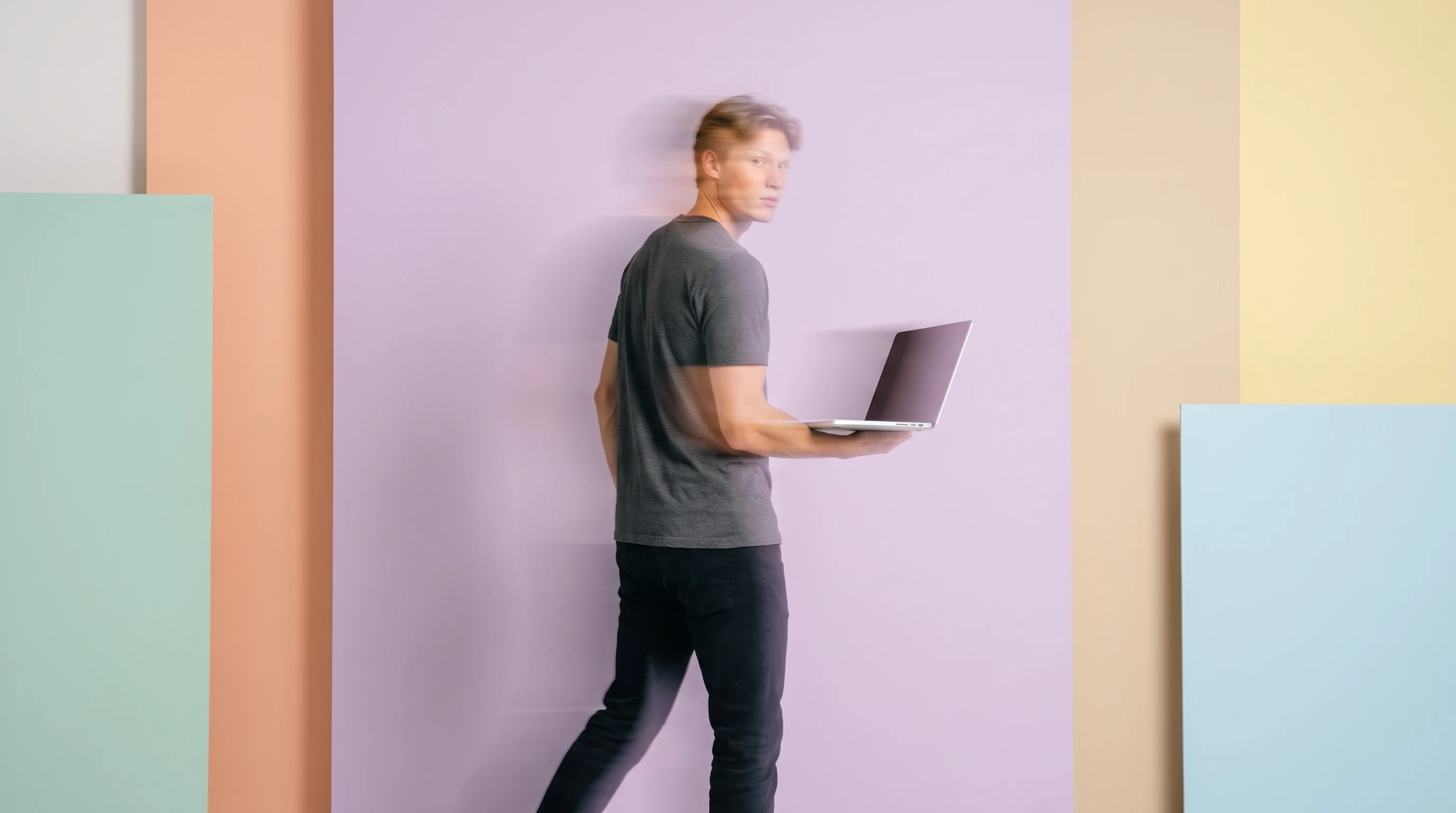 A person mid-step from behind in a lilac backdrop studio, carrying a slim open laptop at hip height, wearing a charcoal crew-neck, glancing back over