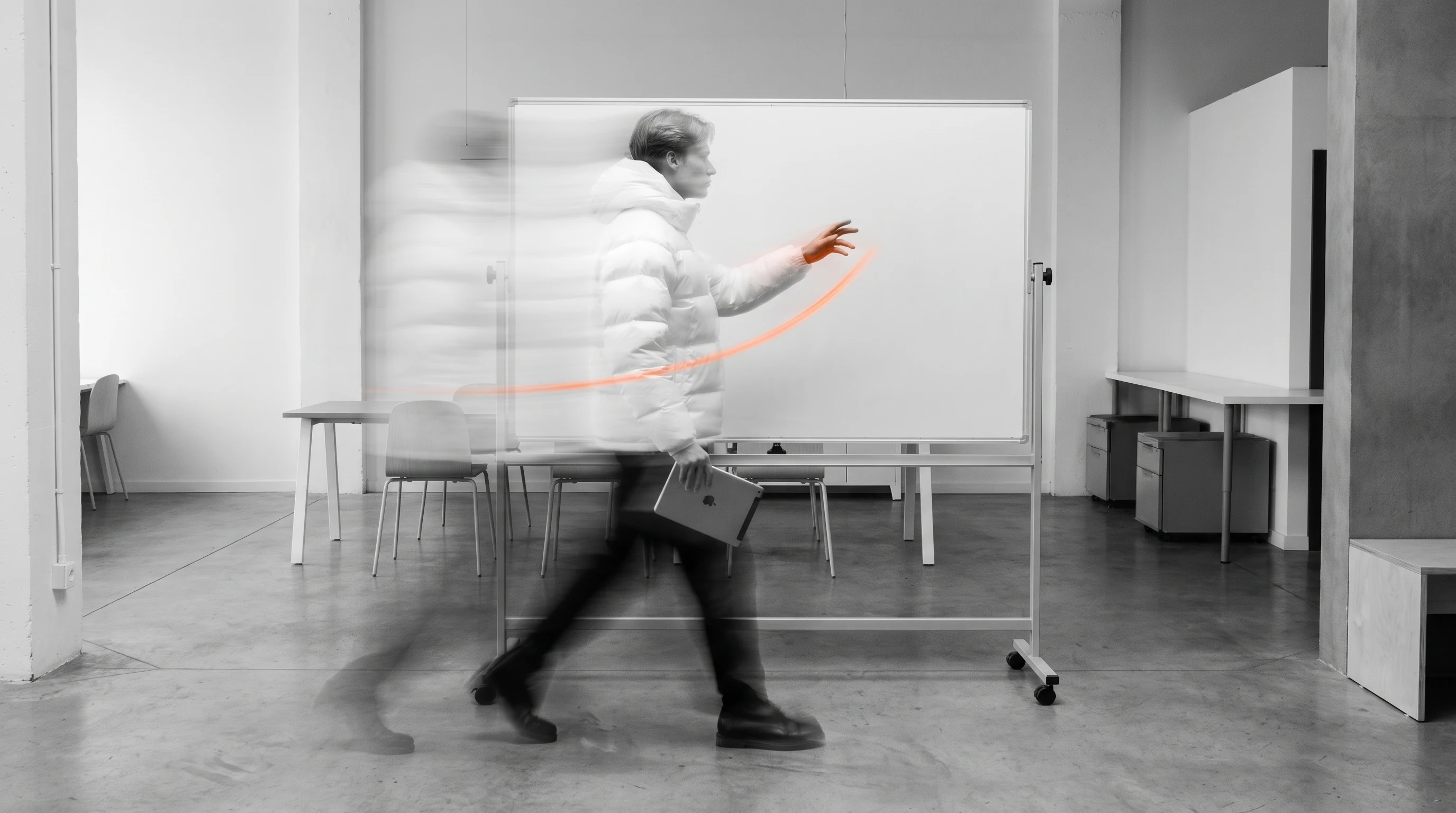 A person mid-step through a clean open workspace, gesturing toward a blank whiteboard with one hand and holding a tablet face-down in the other,