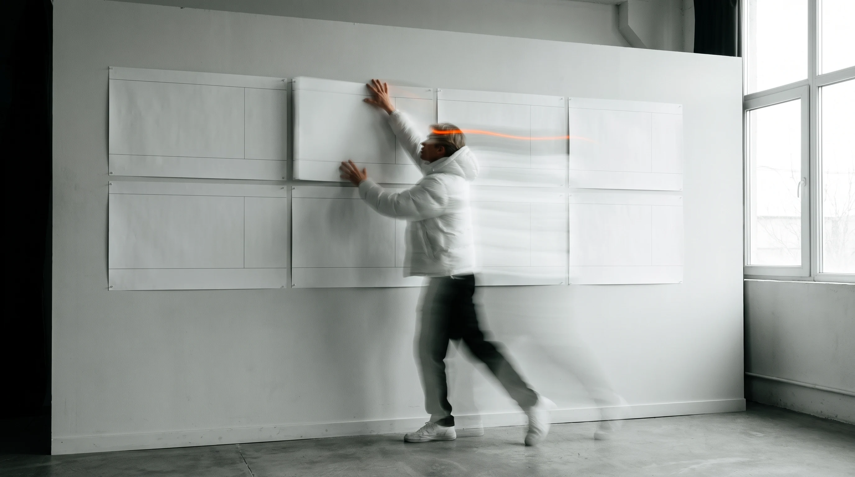 A person mid-step in a wide studio, reaching up to rearrange large blank storyboard panels pinned across a wall, three-quarter framing