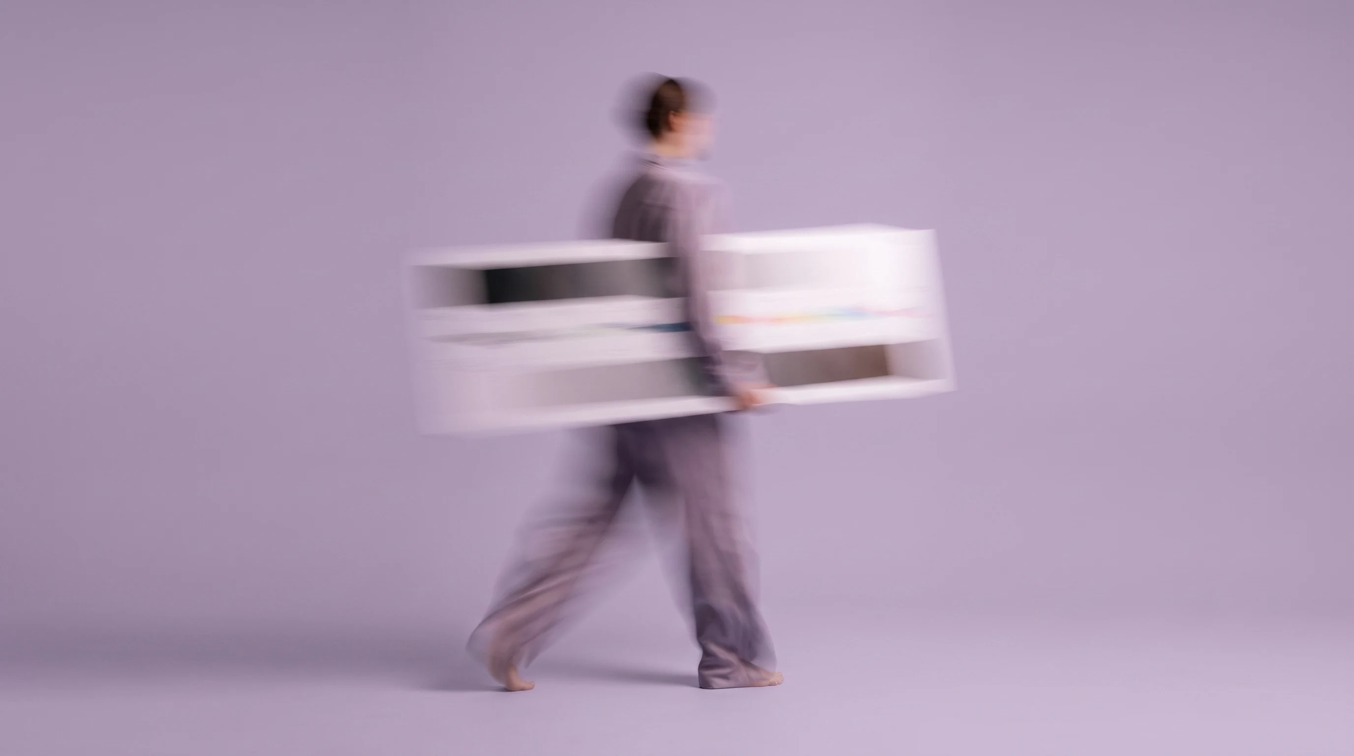 A person mid-step in a lilac studio, holding a large printed aspect-ratio reference card horizontally, framed from behind at waist height