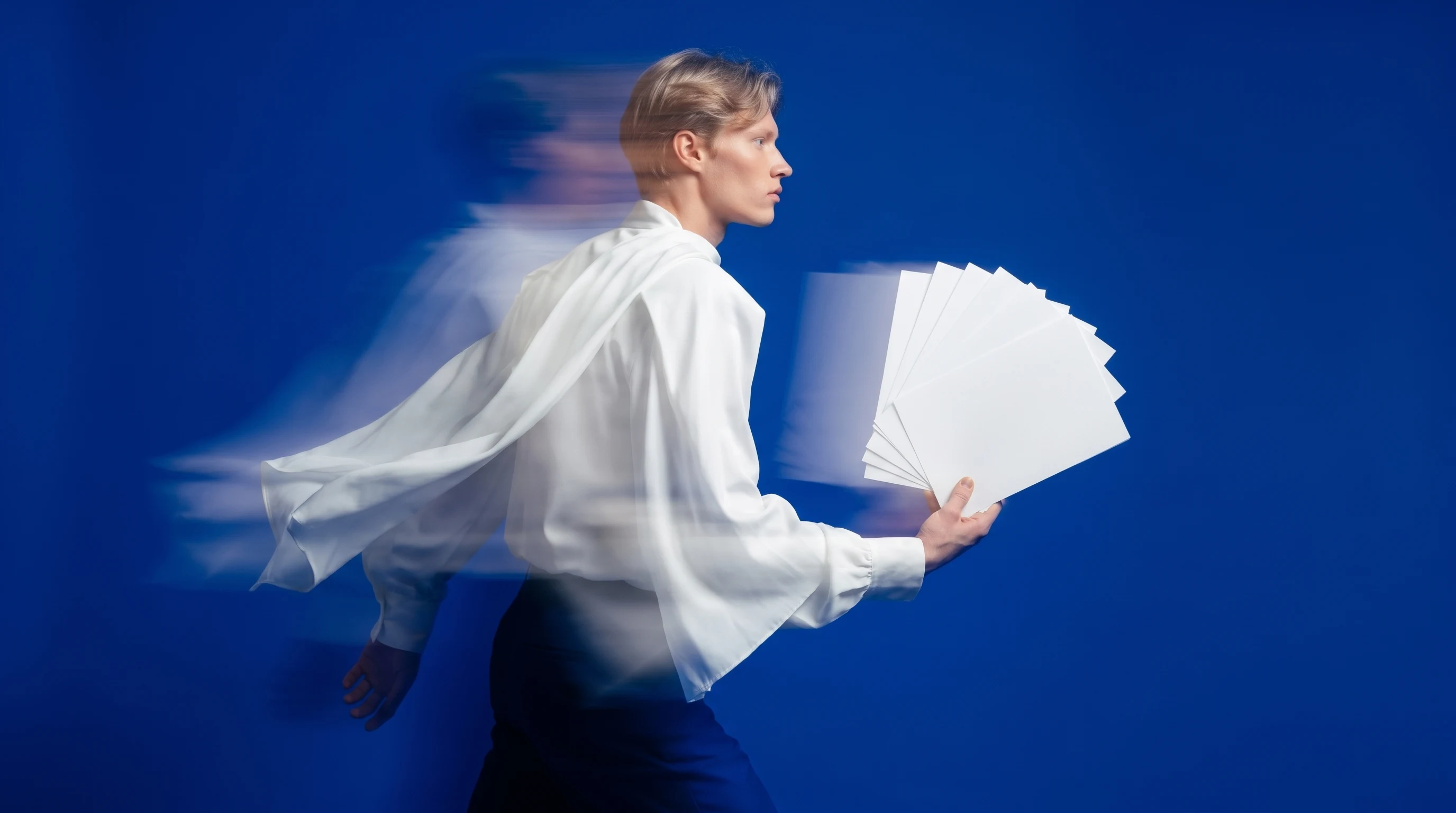 A person mid-step in a cobalt-blue studio, holding a fanned stack of unbranded ad cards, bold white blouse, profile framing