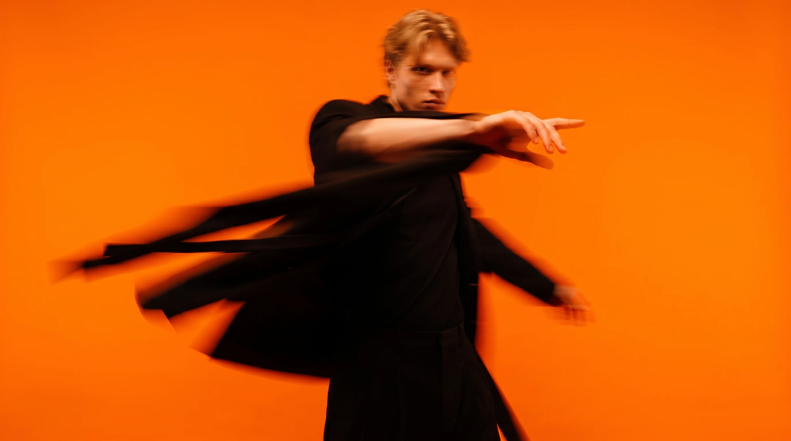 A person mid-spin in a saturated orange studio, arm extended toward camera, black outfit, mid-gesture curating unseen ad creative layouts