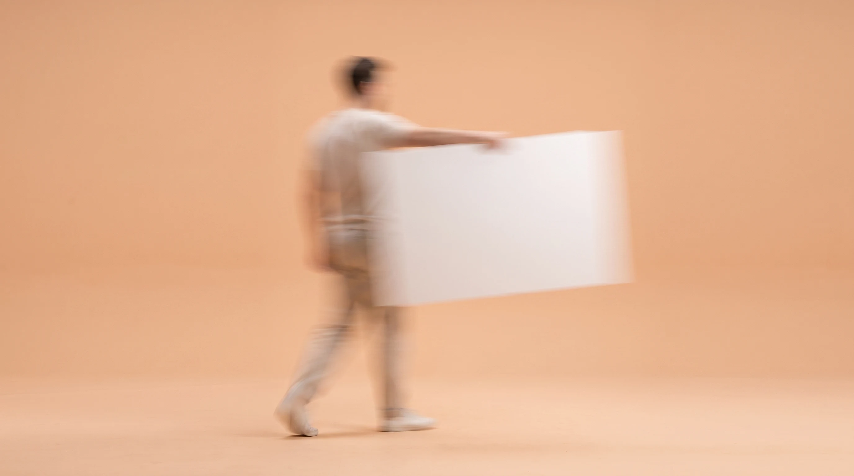 a person mid-step across a peach-backdrop studio, holding a large unbranded storyboard panel at arm's length, framed from behind at a slight angle