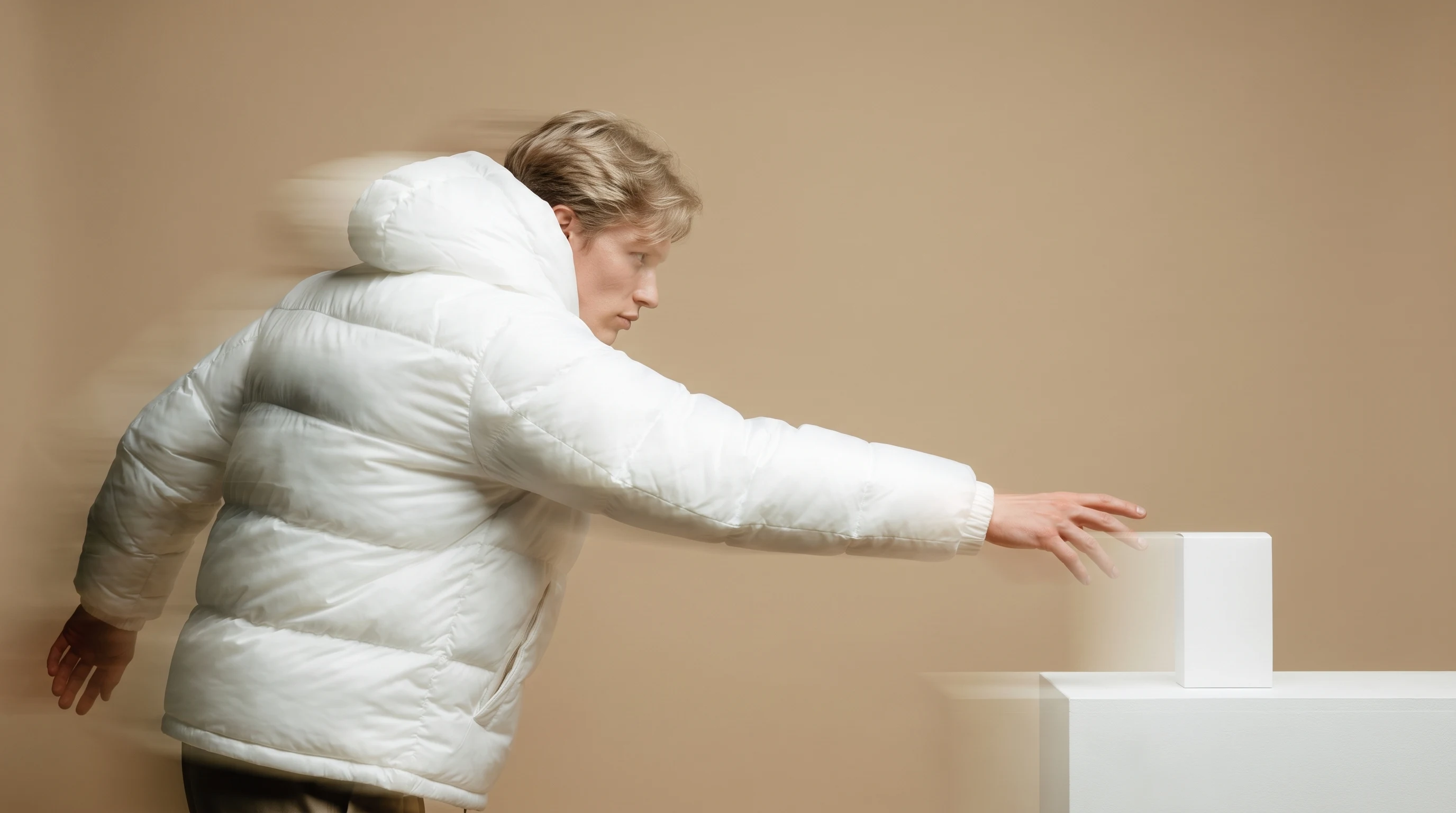 A person mid-reach on a sand-coloured backdrop, extending one arm toward a small unbranded object on a low white plinth, angled three-quarter toward
