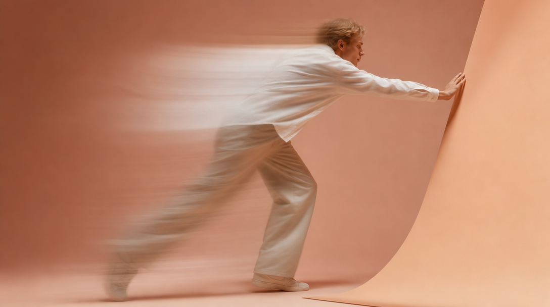 a person mid-step in a peach studio, reaching forward to adjust a plain unbranded paper flat-lay surface, shot from behind in profile, outfit a clean