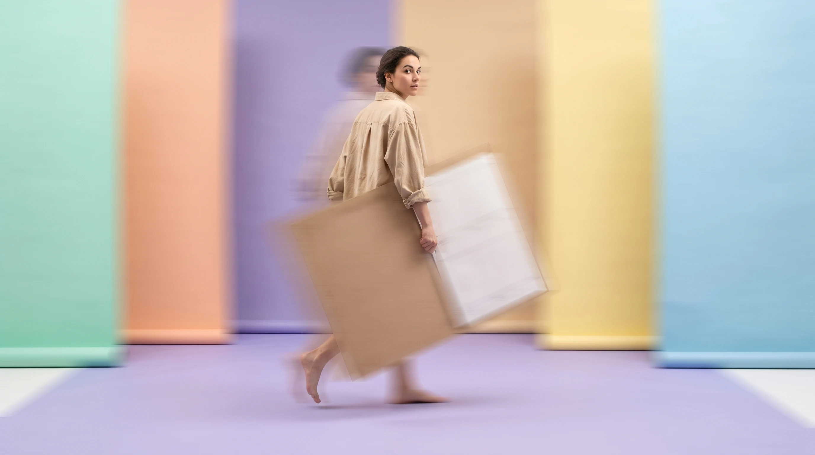 a person mid-step across a lilac studio floor, carrying a large unbranded storyboard panel under one arm, head turned back over shoulder, wearing a