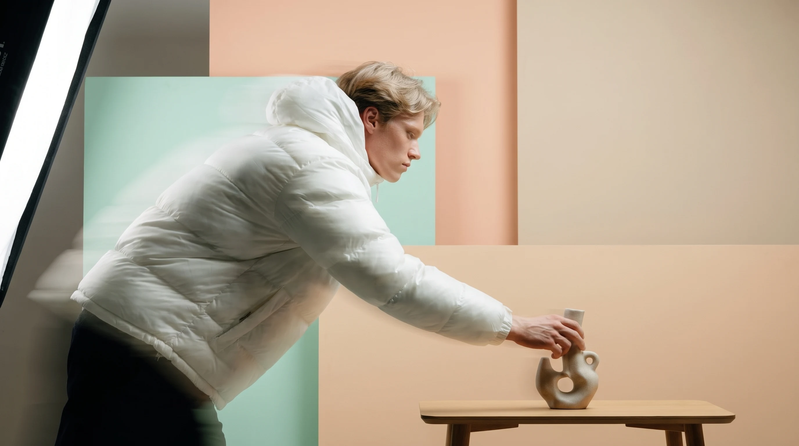 a person mid-reach in a peach-backdrop studio, leaning forward to adjust a small unbranded object on a low table, shot in profile with a softbox edge