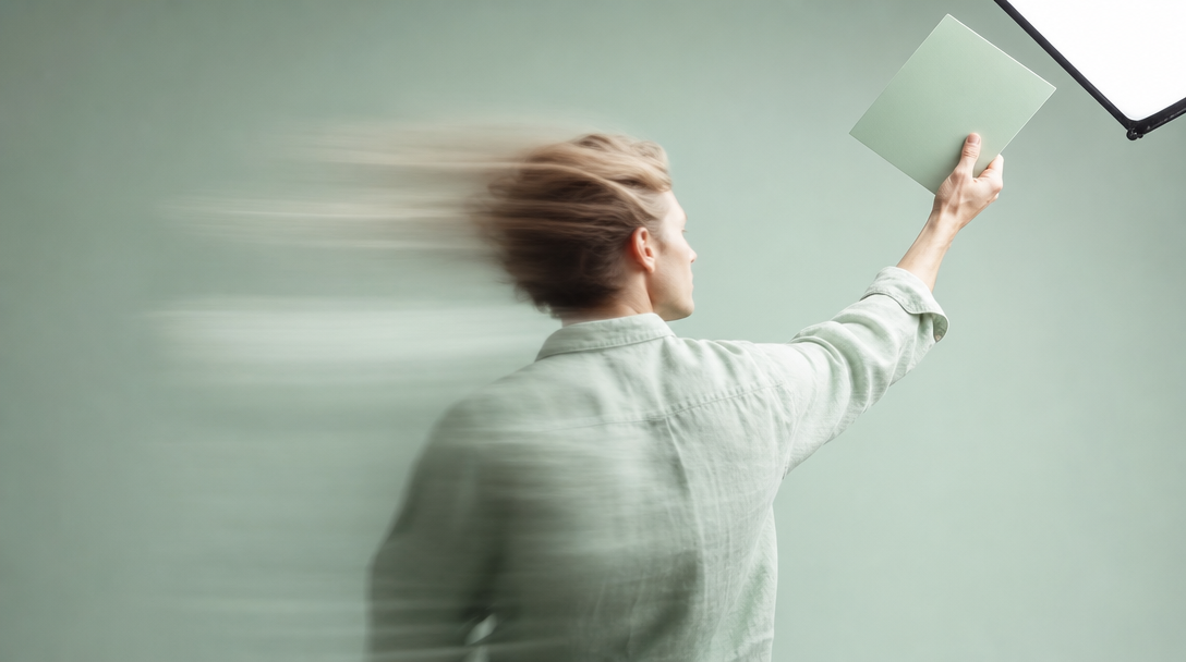 A person in a mint linen shirt mid-turn away from camera, arm raised holding a large unbranded colour-swatch card up toward an overhead softbox just