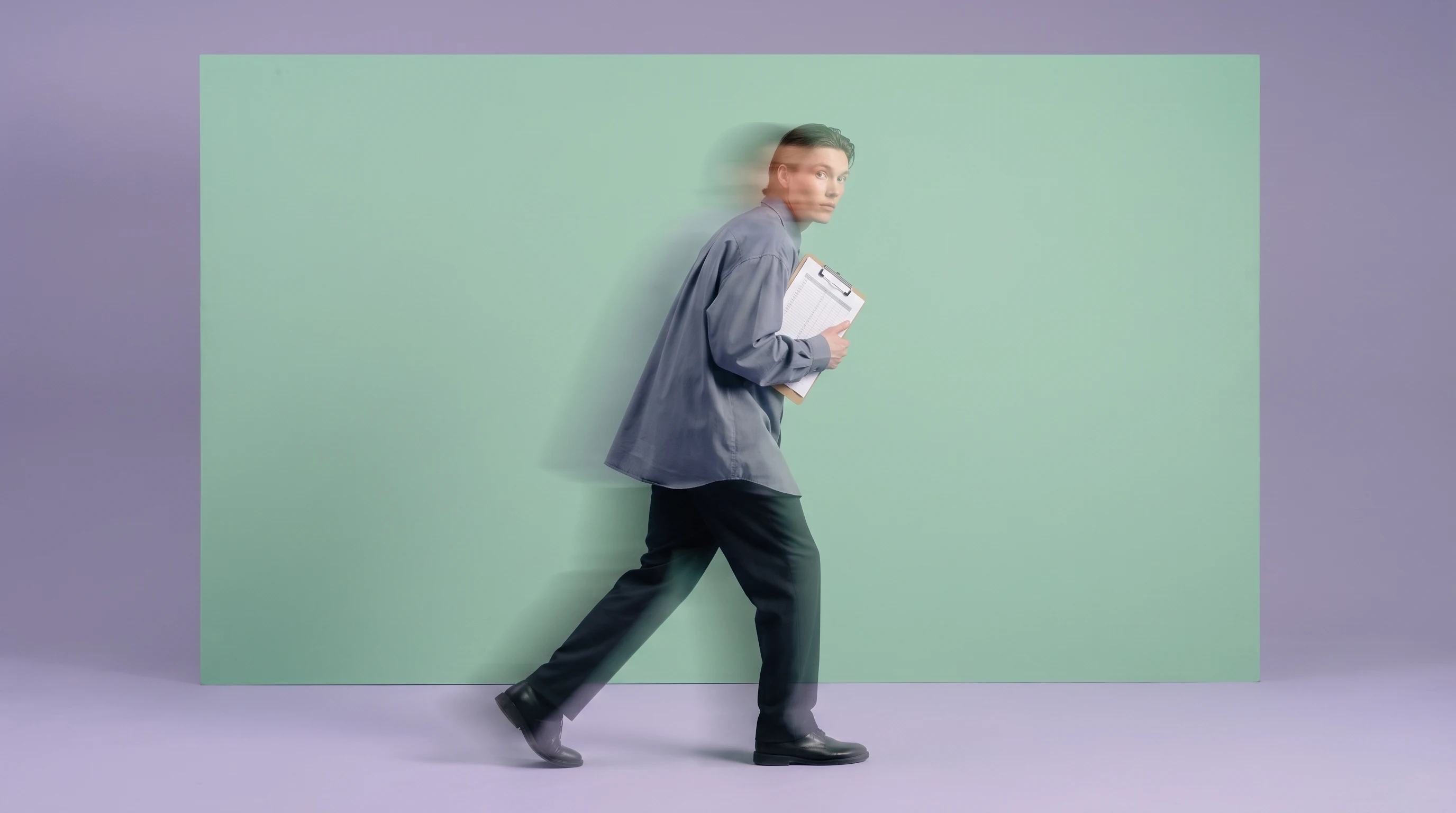 a person mid-step across a lilac studio floor, holding a clipboard close to their chest, glancing back over one shoulder, wearing a slate-grey
