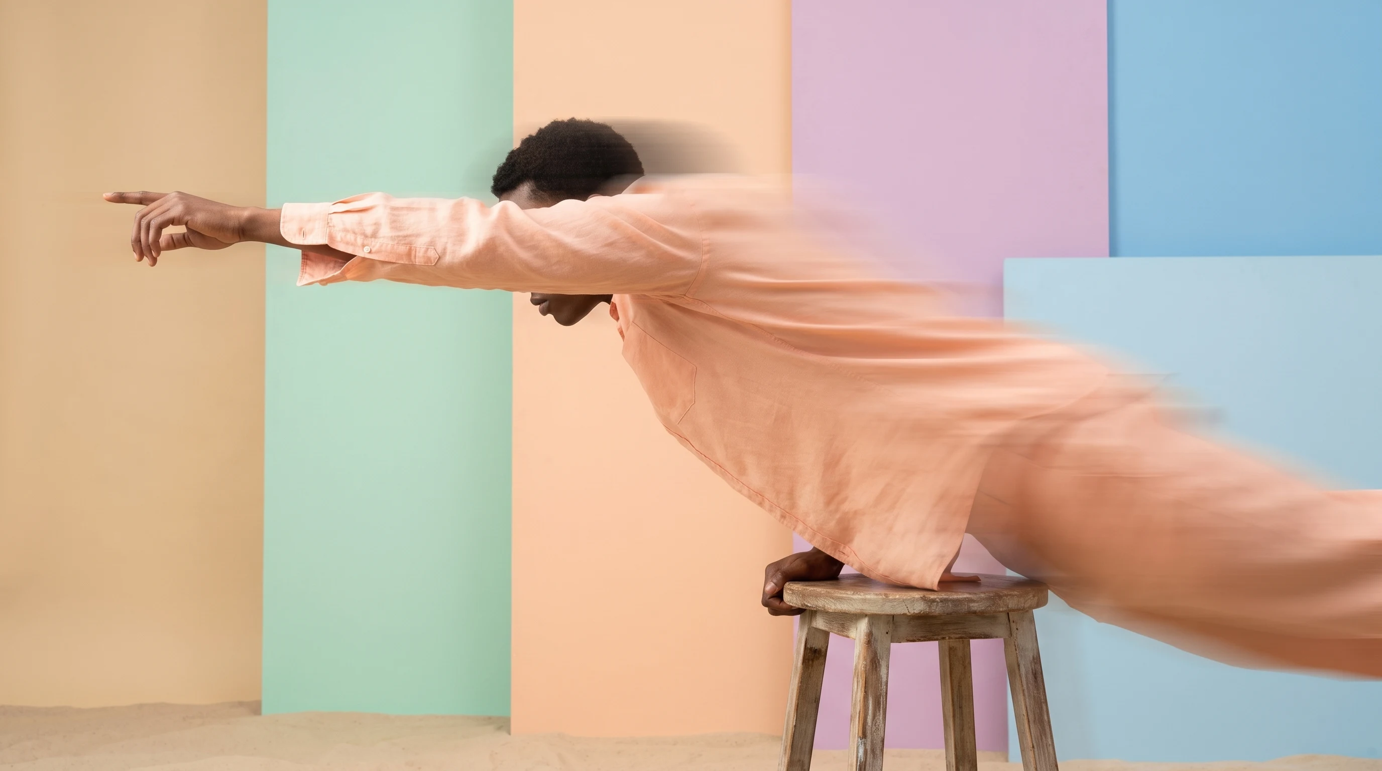 a person in a sand-toned studio mid-lean forward from a stool, arm outstretched pointing off-frame, wearing a soft peach linen shirt, framed in loose