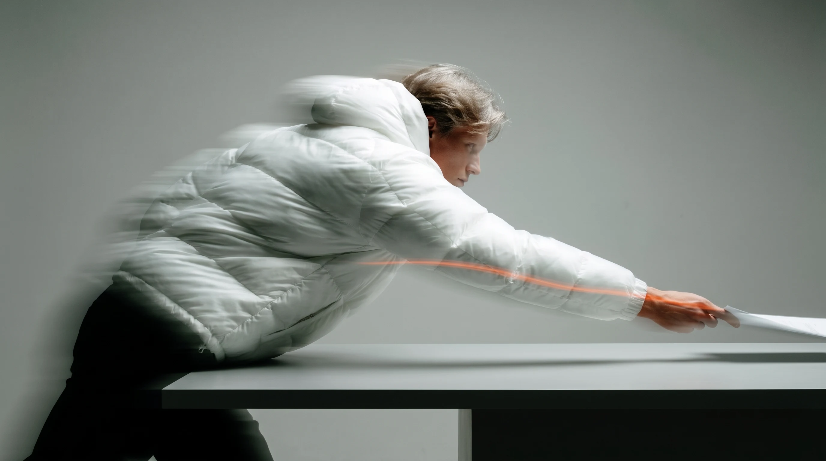 A person mid-reach across a wide desk surface, leaning forward from a standing position, framed in profile, arm extended as if placing or reviewing