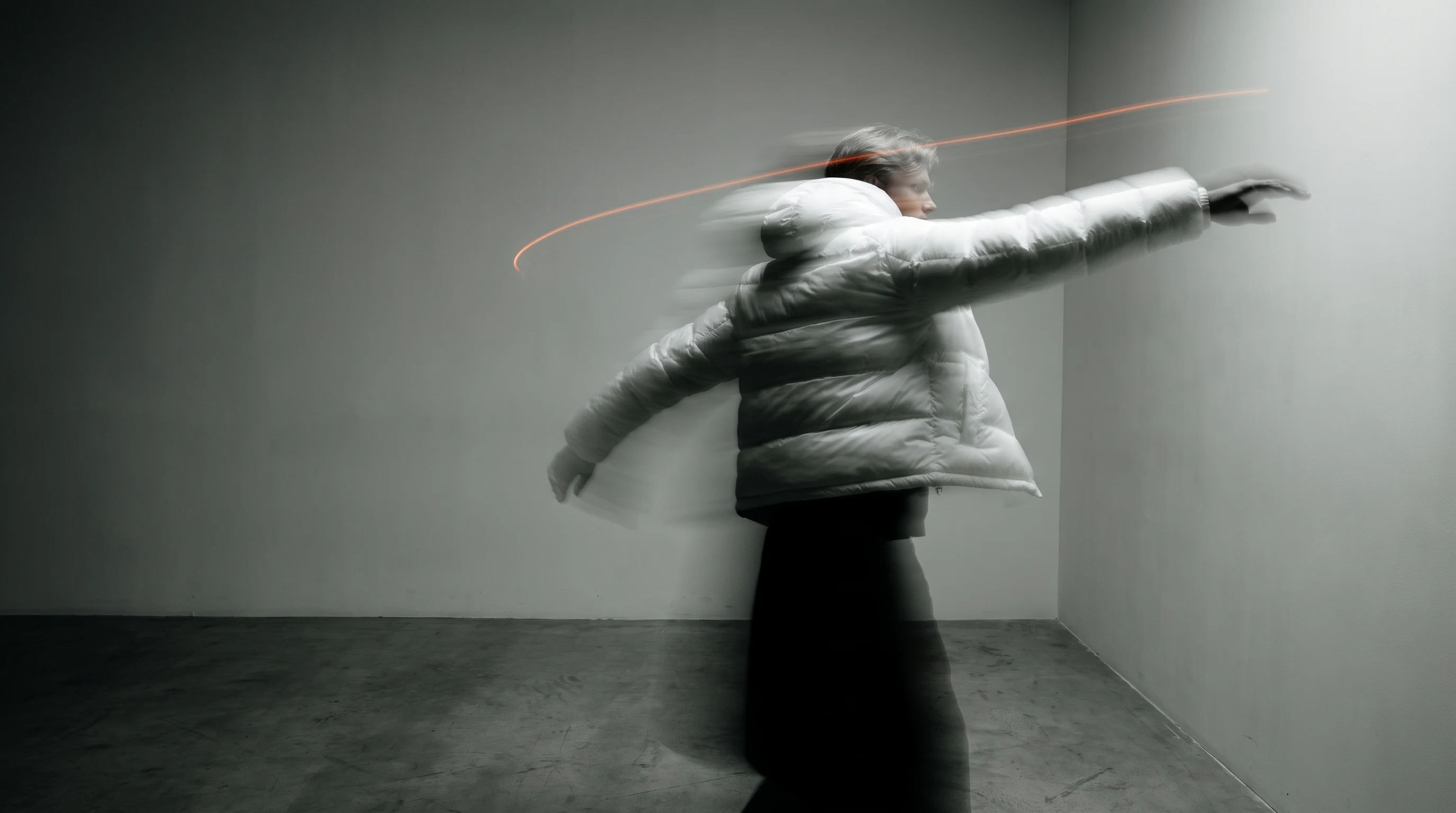 A person mid-turn in a minimal studio space, one arm extended gesturing toward an unseen wall, framed three-quarter, as if mid-pitch presenting a