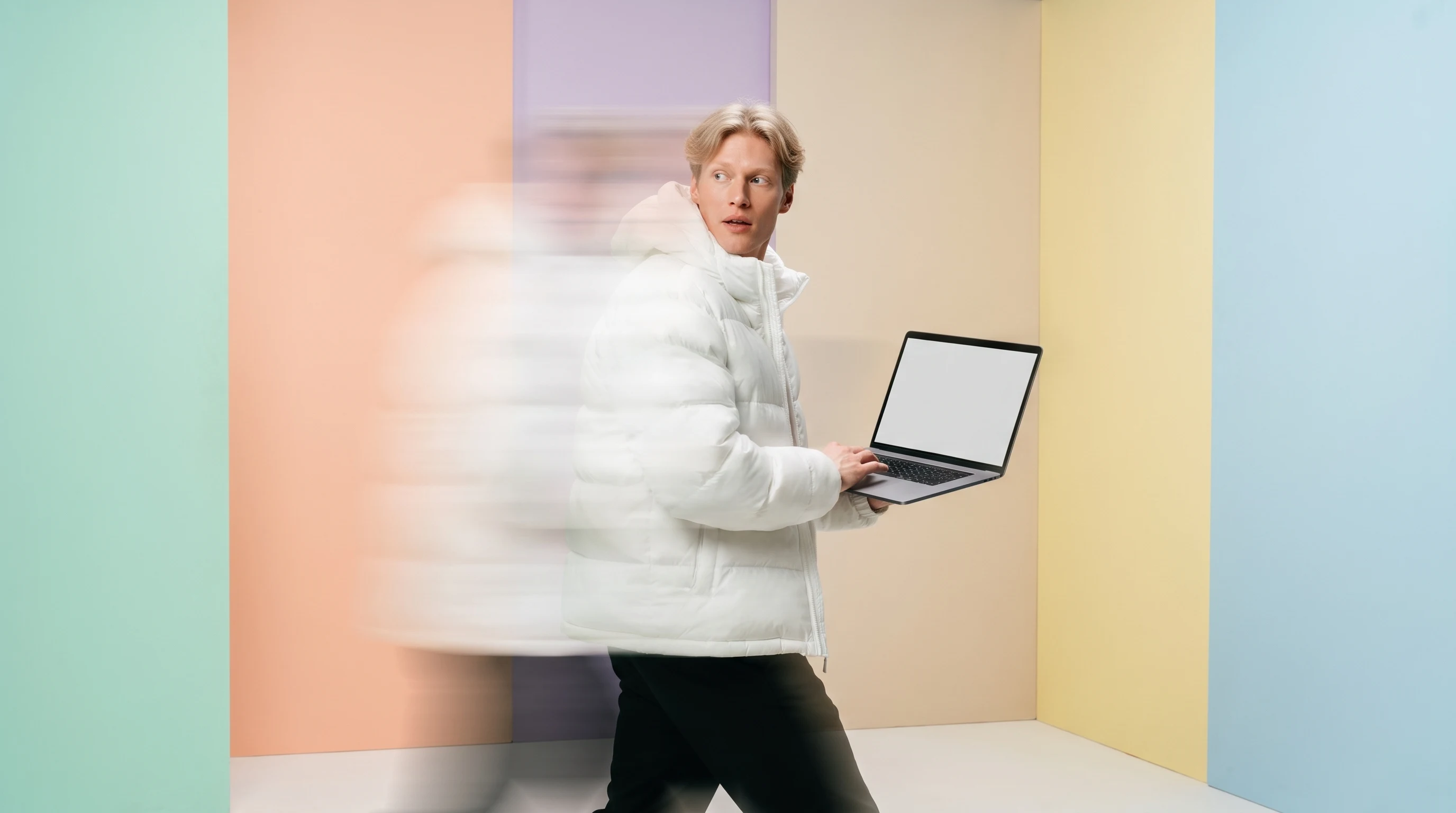 a person mid-step in a mint studio, carrying a blank open laptop at hip height, glancing back over one shoulder as if mid-briefing walkthrough