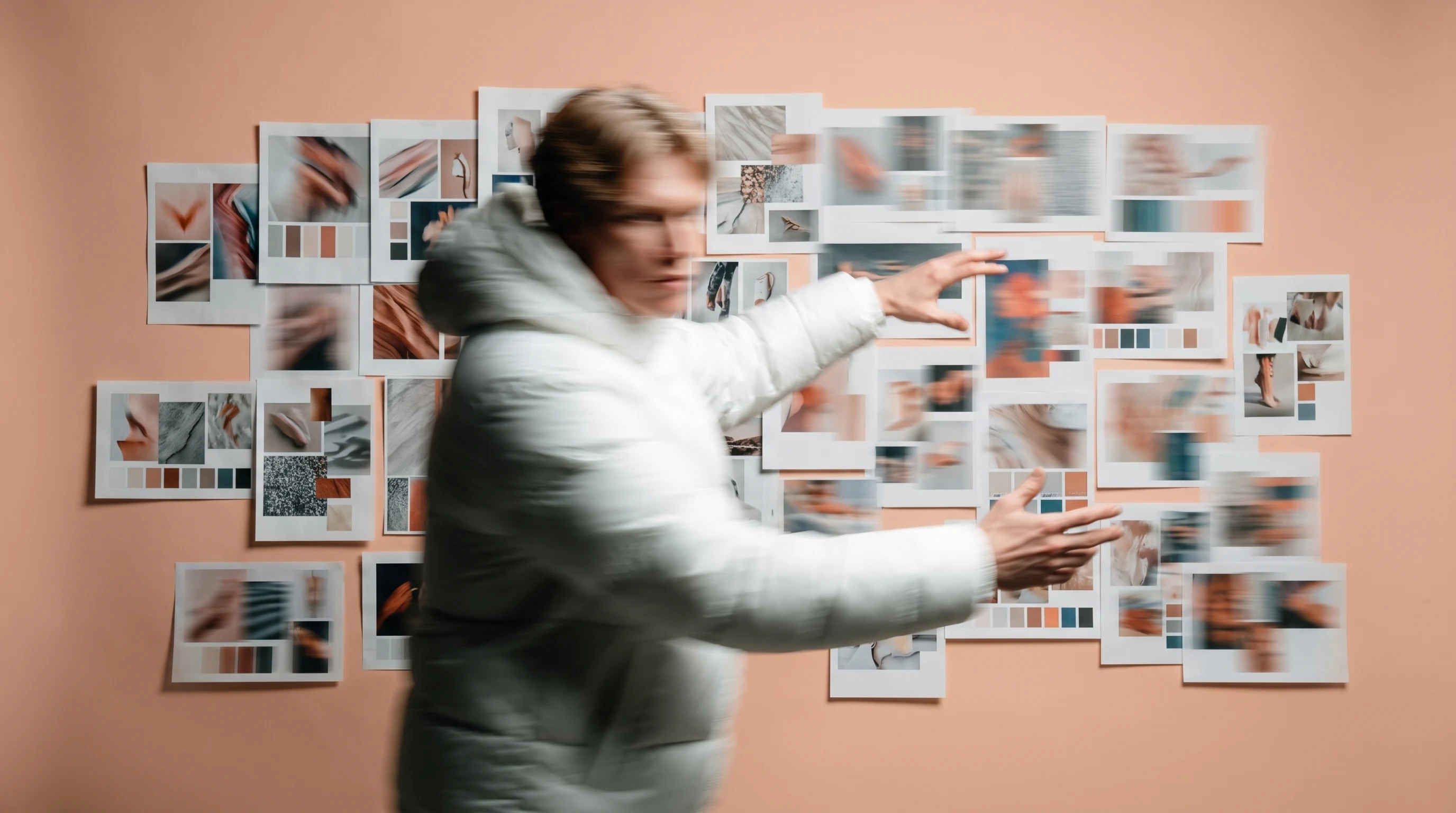 a person mid-turn in a peach studio, arms gesturing outward as if presenting a wall of unbranded mood-board printouts pinned behind them