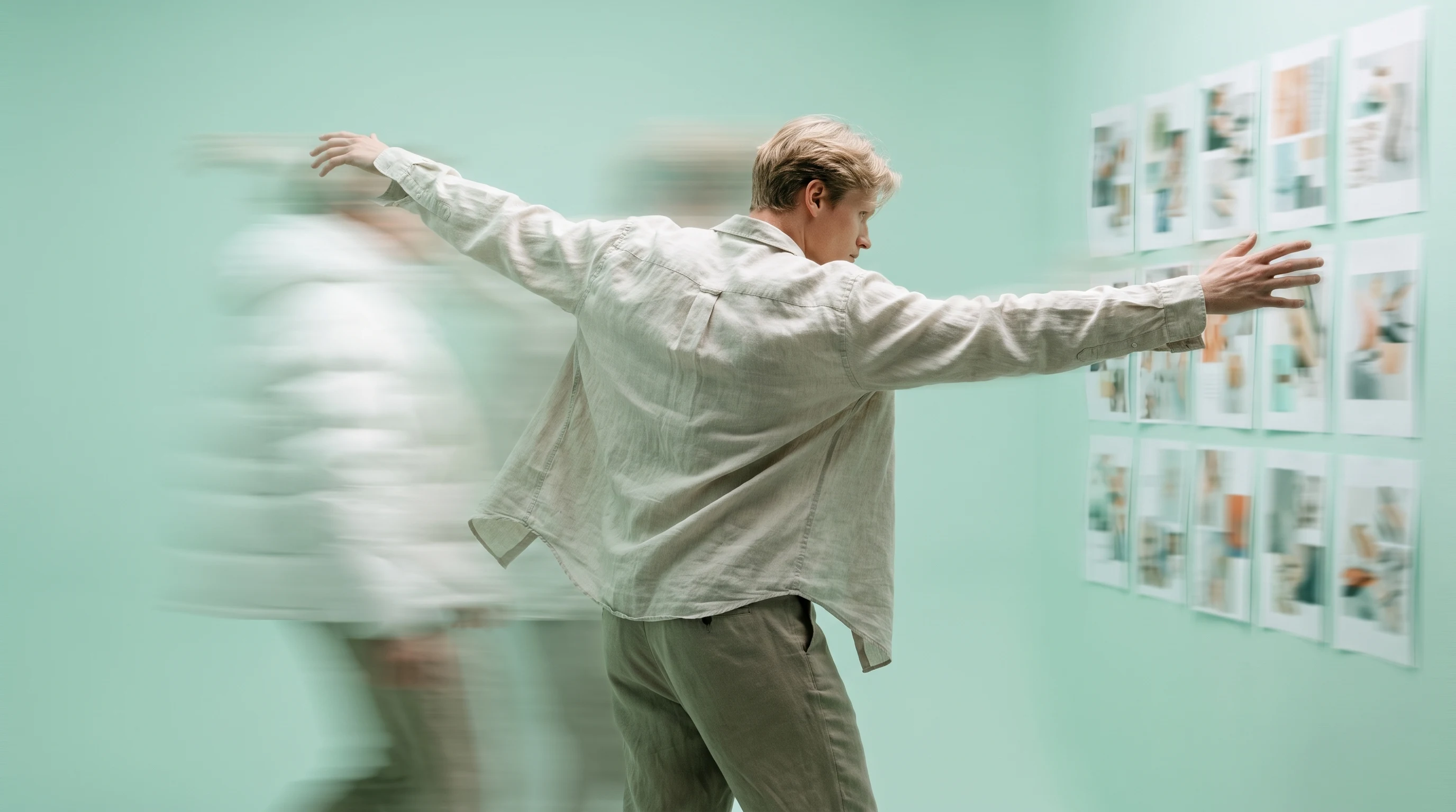 A person mid-turn in a mint-green studio, arms sweeping outward gesturing toward an off-frame wall of unbranded mood-board prints, three-quarter