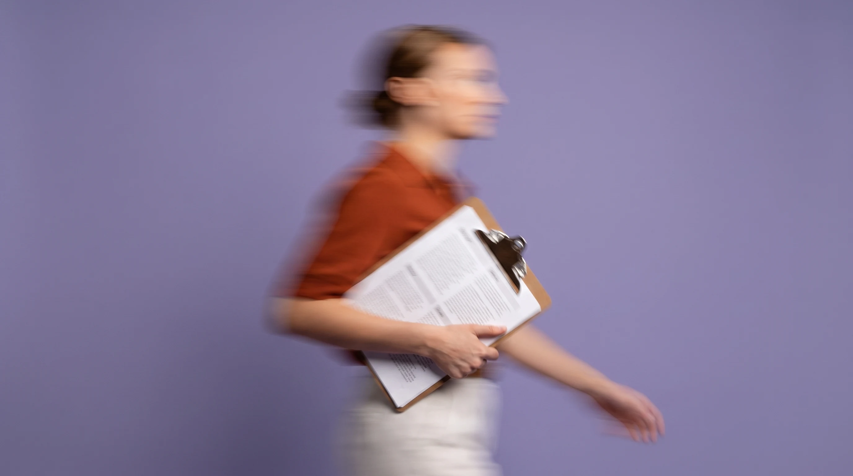 A person mid-step across a lilac studio backdrop, holding a clipboard of printed pages to their chest, wearing a rust-orange top, framed from the