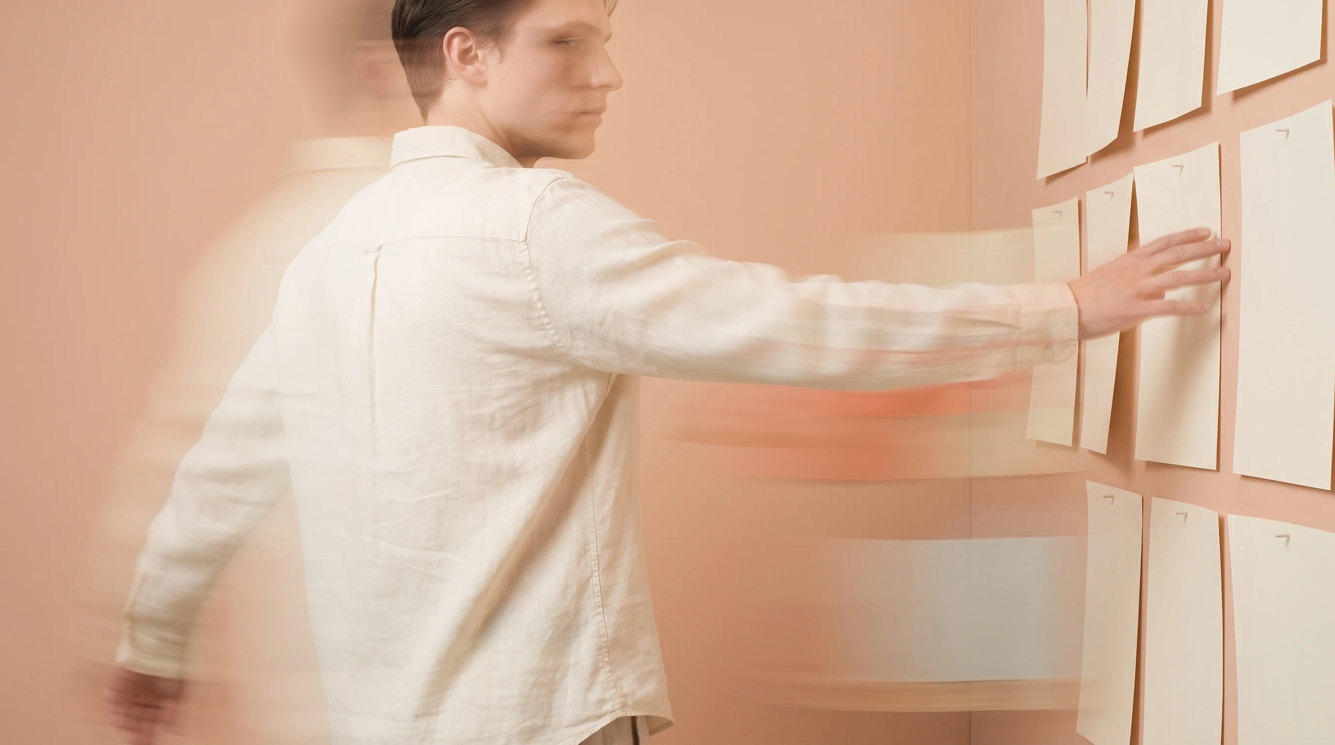 A person mid-turn in a peach studio, arm extended toward pinned blank papers on a wall, three-quarter framing, cream linen shirt
