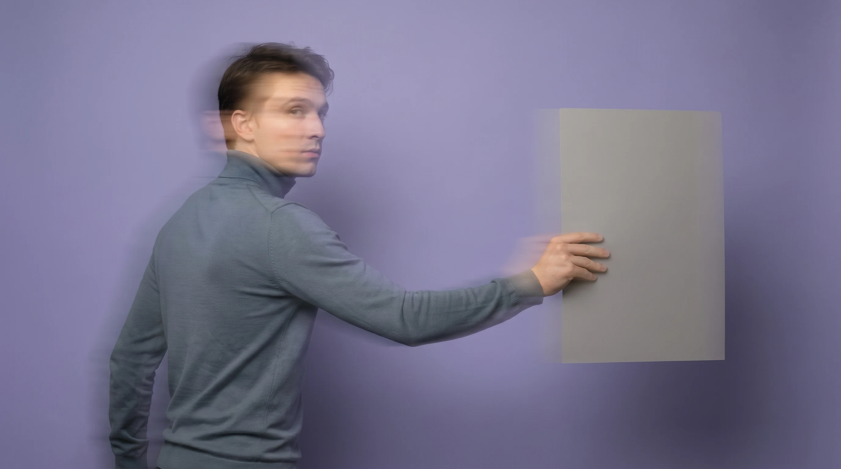A person mid-step in a lilac-backdrop studio, holding a large unbranded reference card at arm's length and glancing back over one shoulder, wearing a