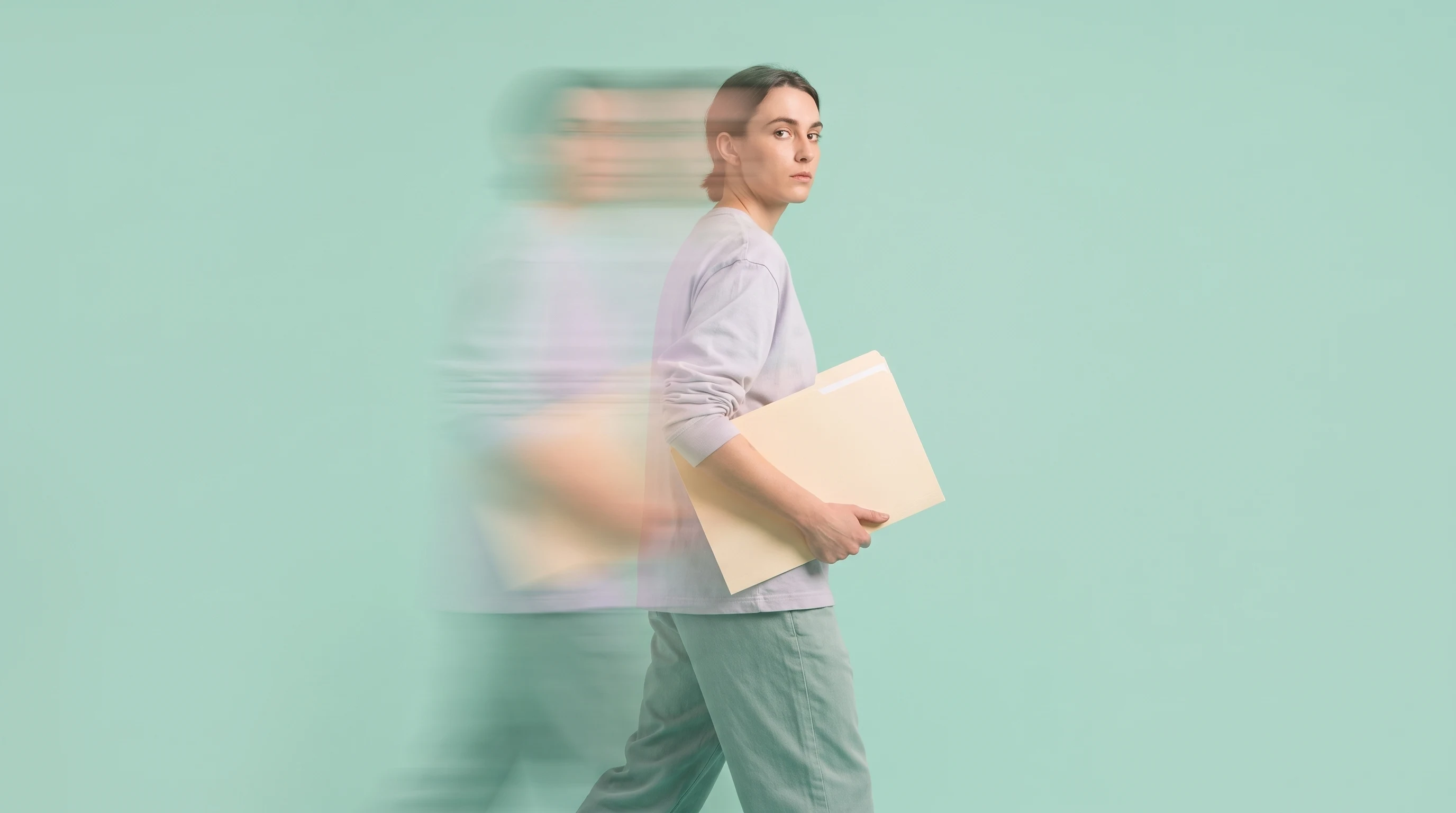 a person mid-step in a mint studio holding a blank unbranded folder, glancing back over one shoulder toward camera, profile framing