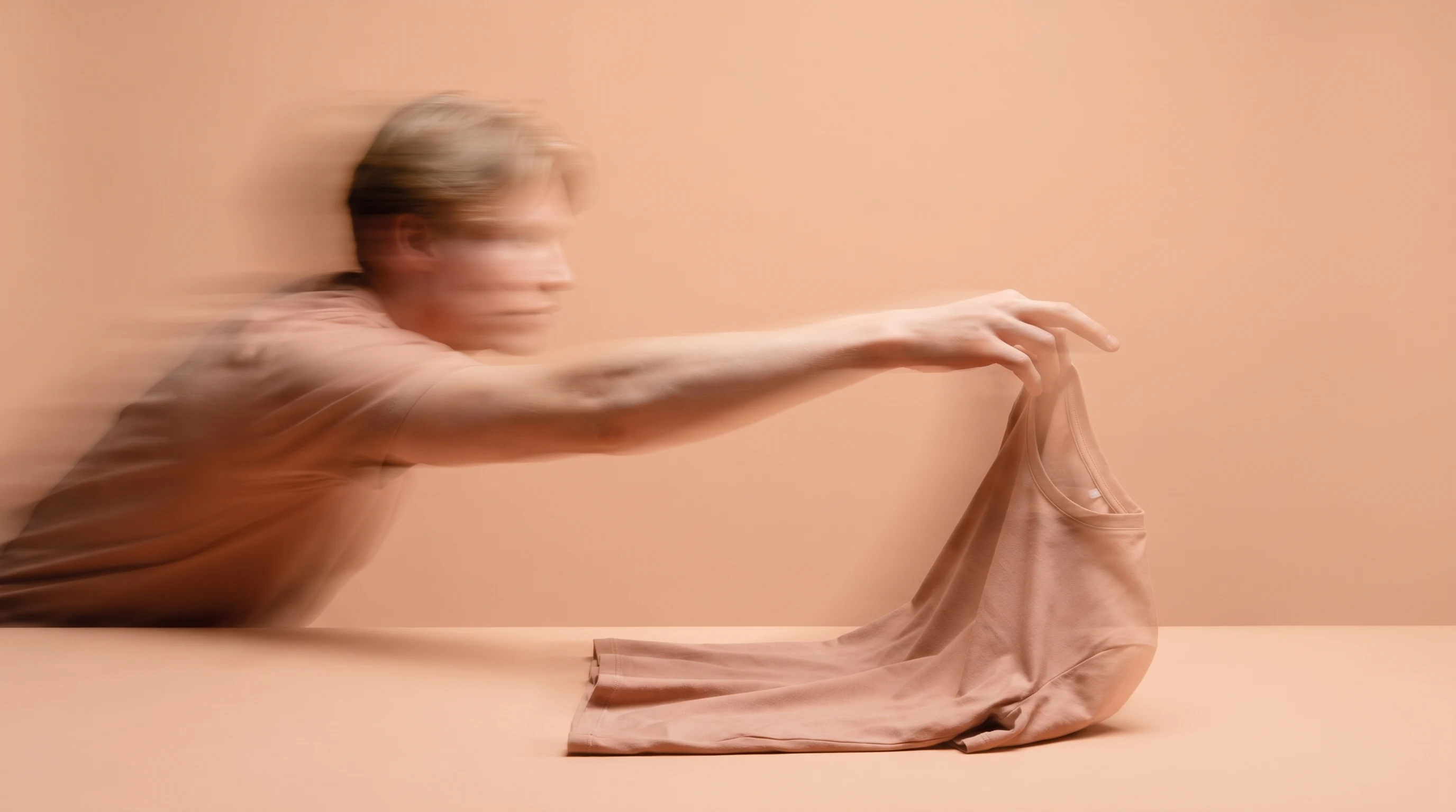 a person mid-reach in a peach studio, draping a plain unbranded folded top across a flat lay surface, profile framing with one arm extended toward
