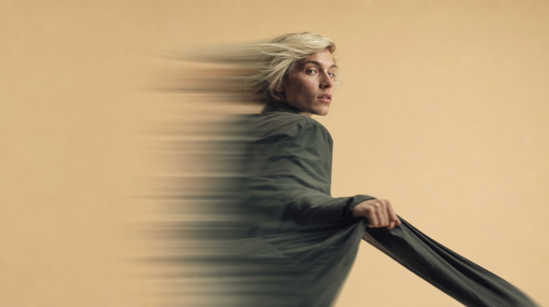 A person mid-turn on a sand studio backdrop, holding a plain unbranded garment at arm's length and glancing back over one shoulder, profile framing