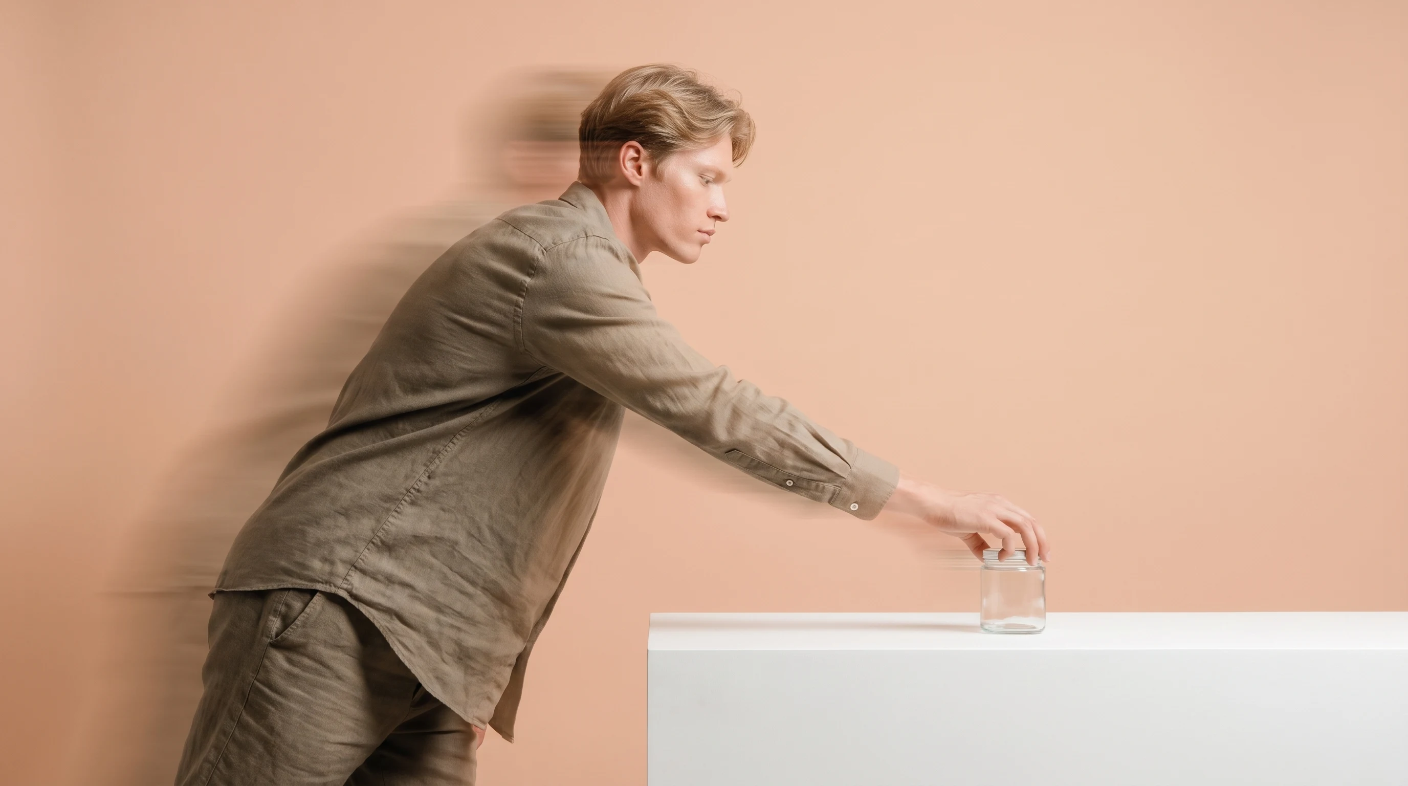A person mid-reach on a peach backdrop, adjusting a small unbranded jar on a minimal white surface, profile framing, different outfit from hero