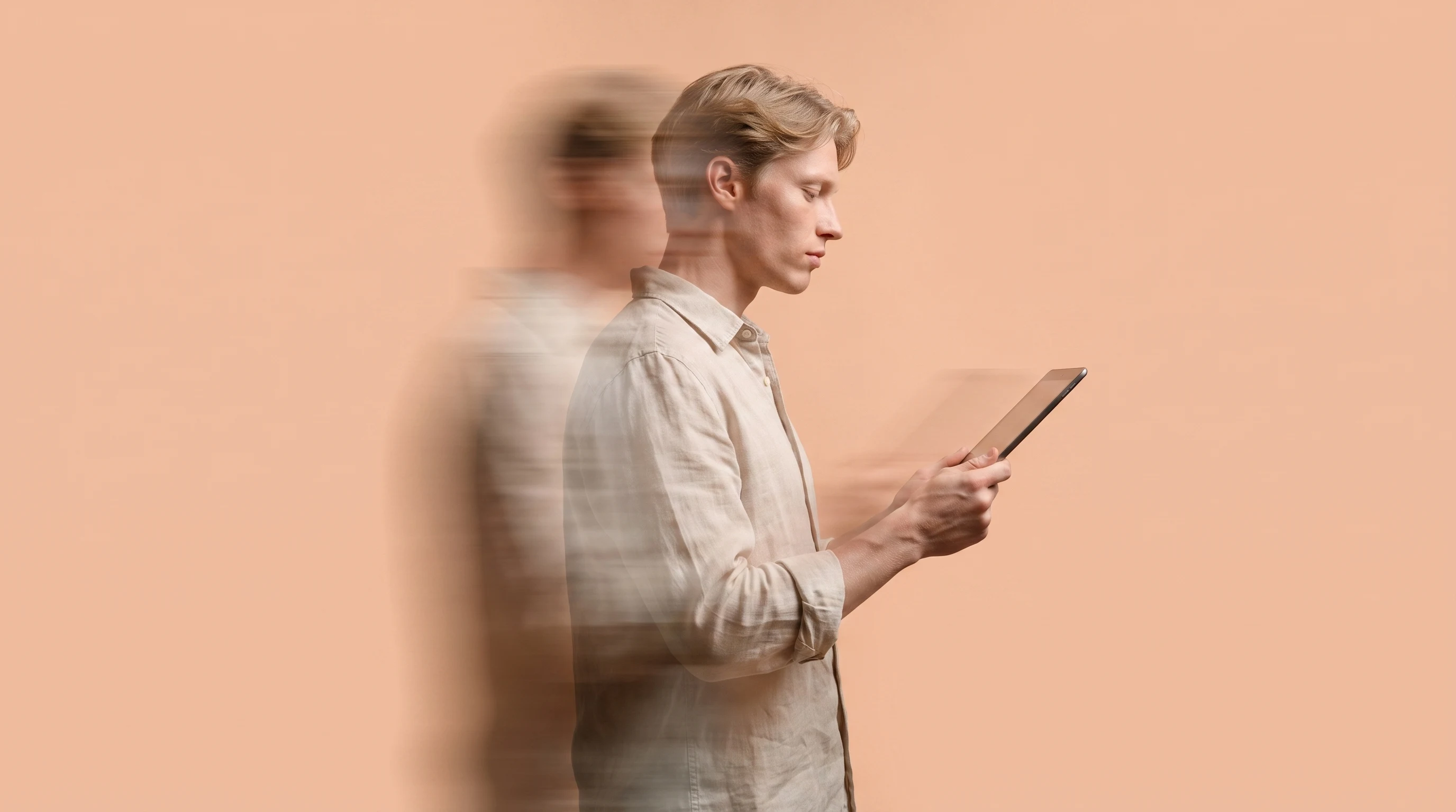 a person mid-turn in a peach studio holding a tablet at chest height, glancing down at the screen with a considered expression, relaxed linen shirt,