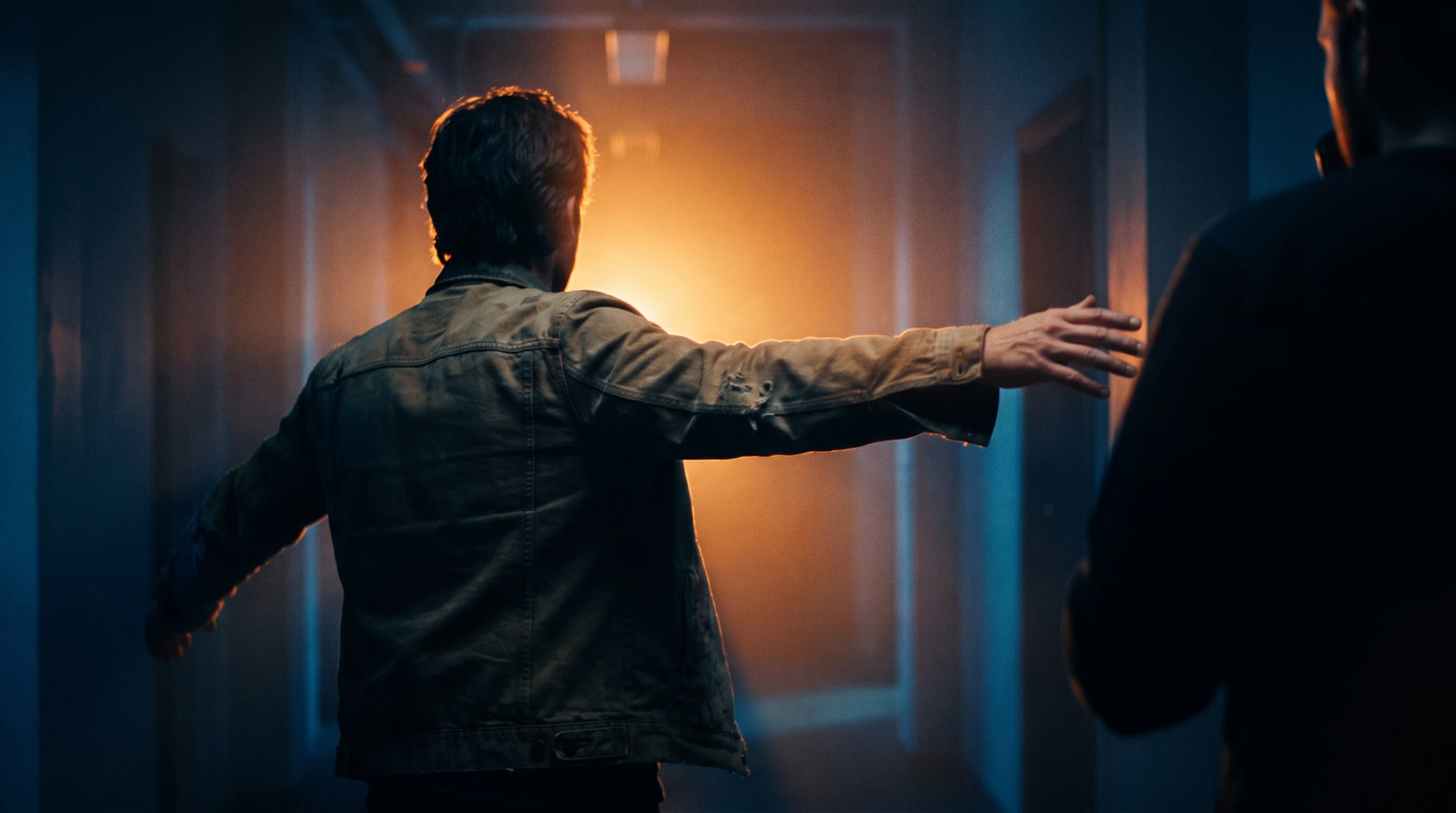 A creative director mid-spin in a darkened studio corridor, arm outstretched to cue an unseen shot, framed low from behind with warm backlight