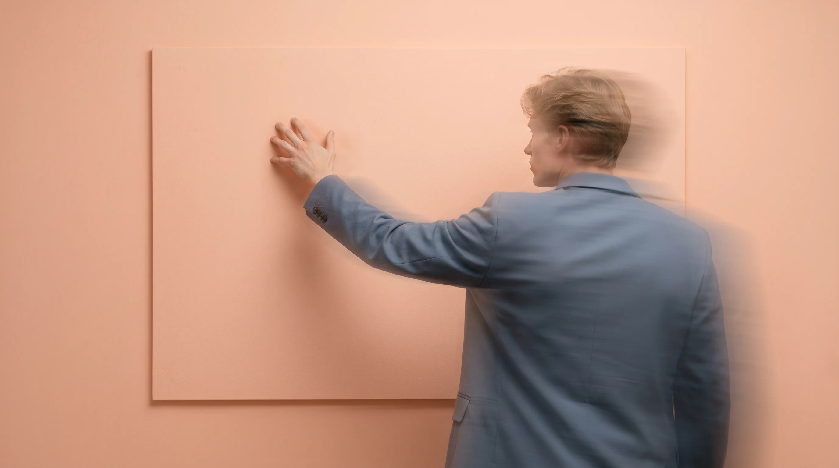 a person mid-reach toward a blank wall-mounted mood board in a peach studio, wearing a slate-blue jacket, framed from slightly behind at a