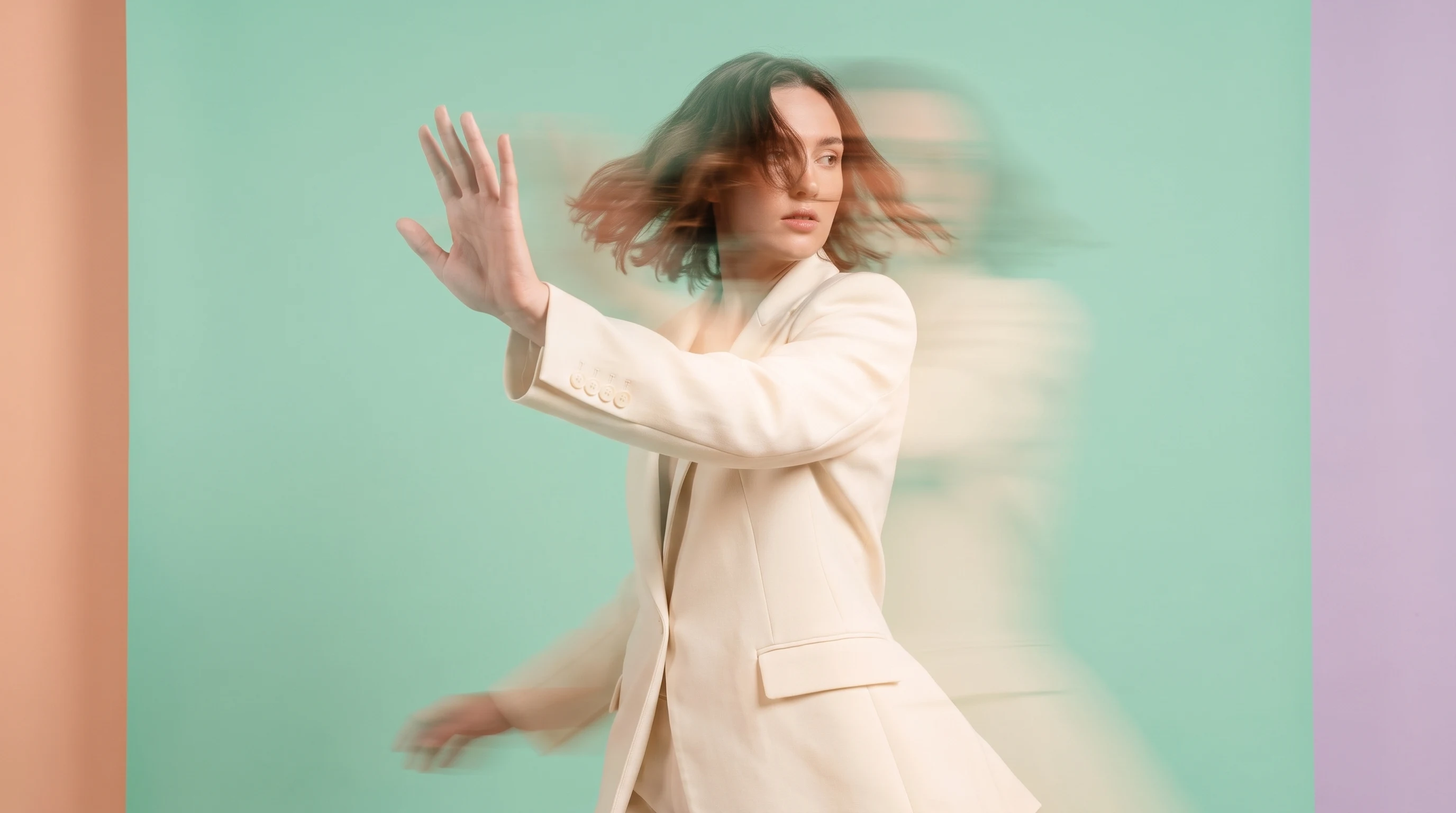 a person mid-turn in a mint-green studio, one hand raised open-palmed in a decisive gesture, wearing a structured cream blazer, framed three-quarter