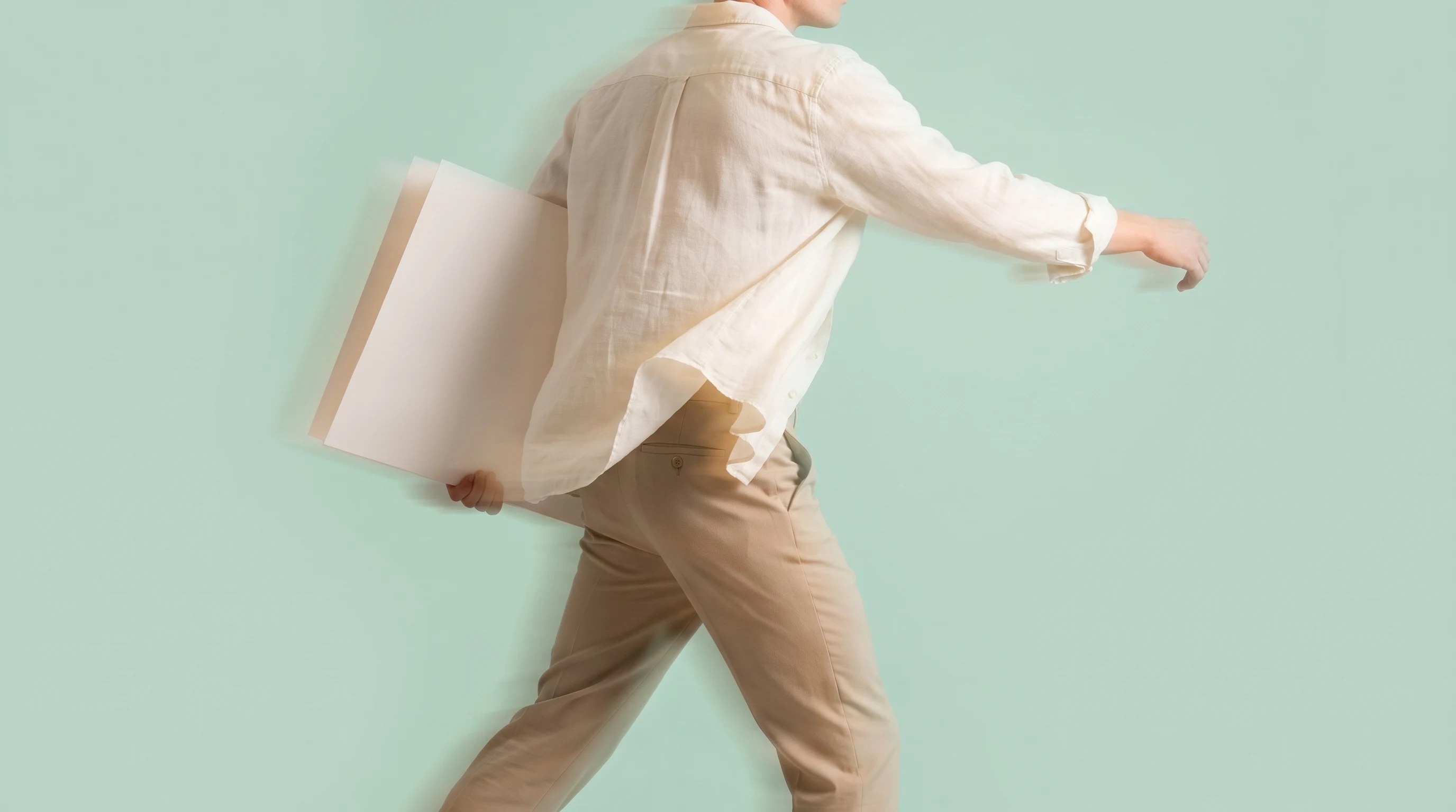 A person mid-step in a mint studio, carrying a large unbranded print under one arm and reaching forward, cropped at mid-thigh, shot from slightly