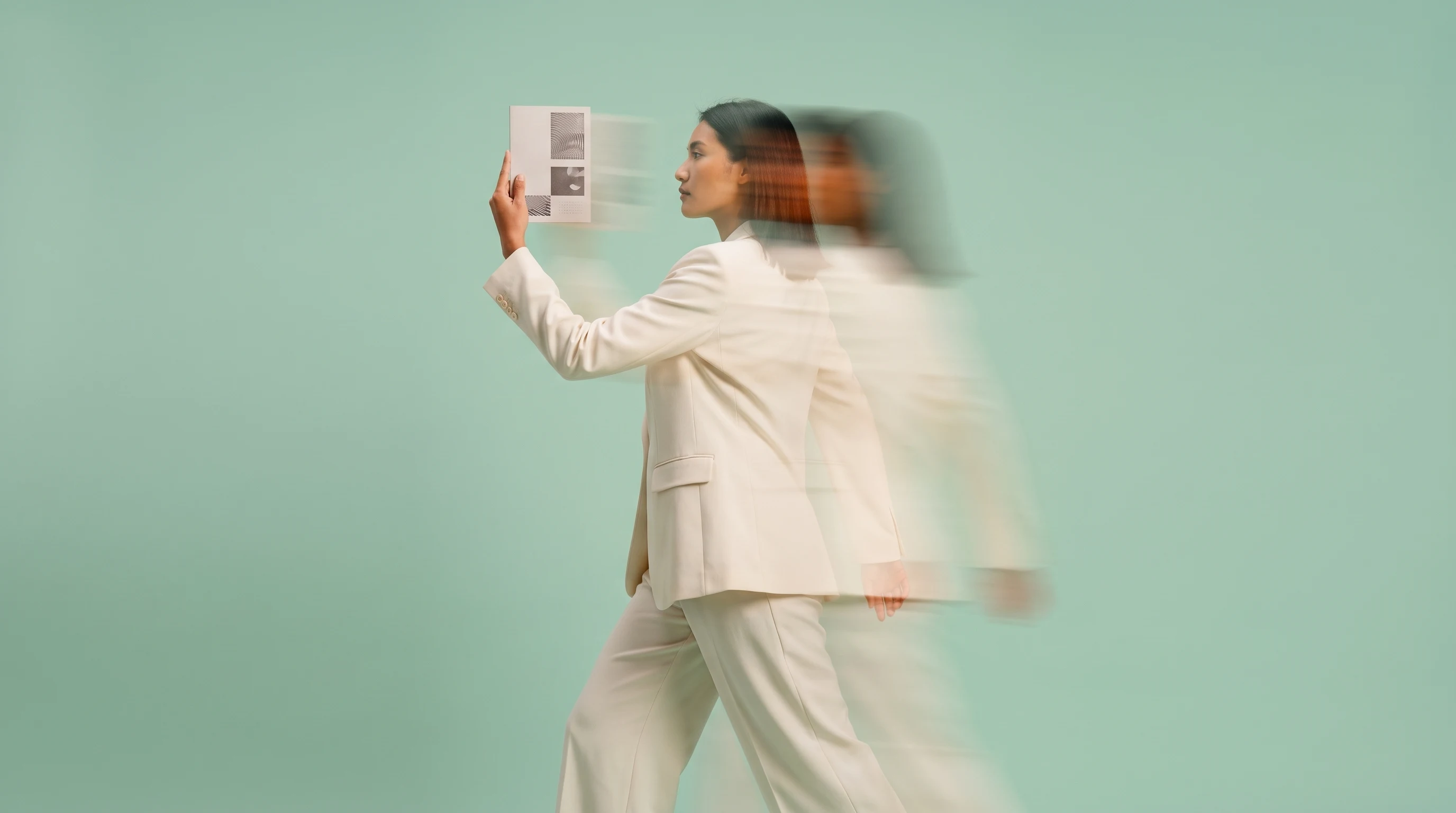 A person mid-turn in a mint studio, one arm raised holding an unbranded printed agenda card at eye level, side-on framing, smart blazer and wide-leg