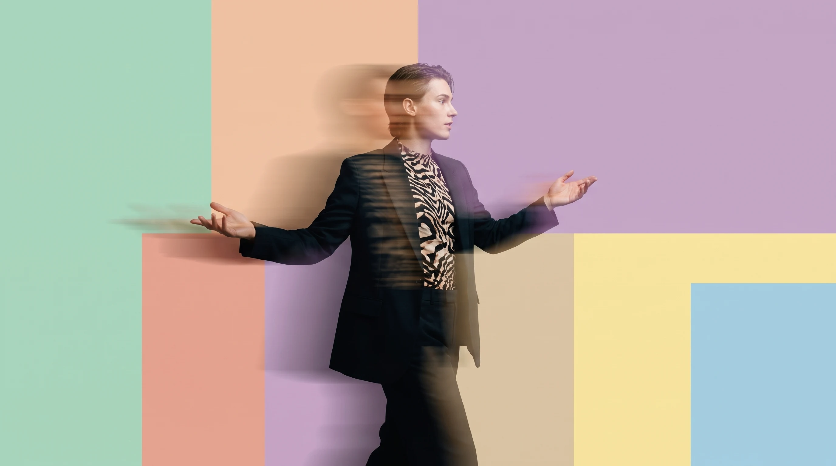 A person mid-stride in a peach studio, arms outstretched mid-gesture as if addressing a conference audience, three-quarter framing, blazer over a