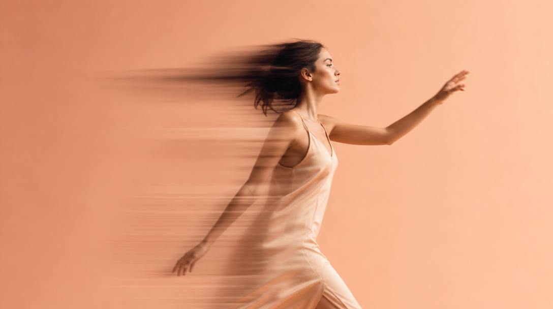 A woman mid-stride through a peach-backdrop studio wearing a sleeveless unbranded slip dress, one hand raised gesturing toward an off-frame