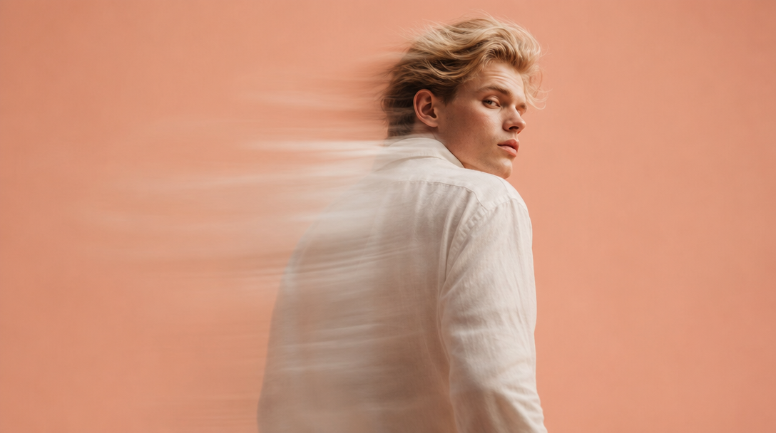 A man mid-step from behind on a peach backdrop, glancing back over his left shoulder with a controlled gaze, wearing a structured linen shirt,