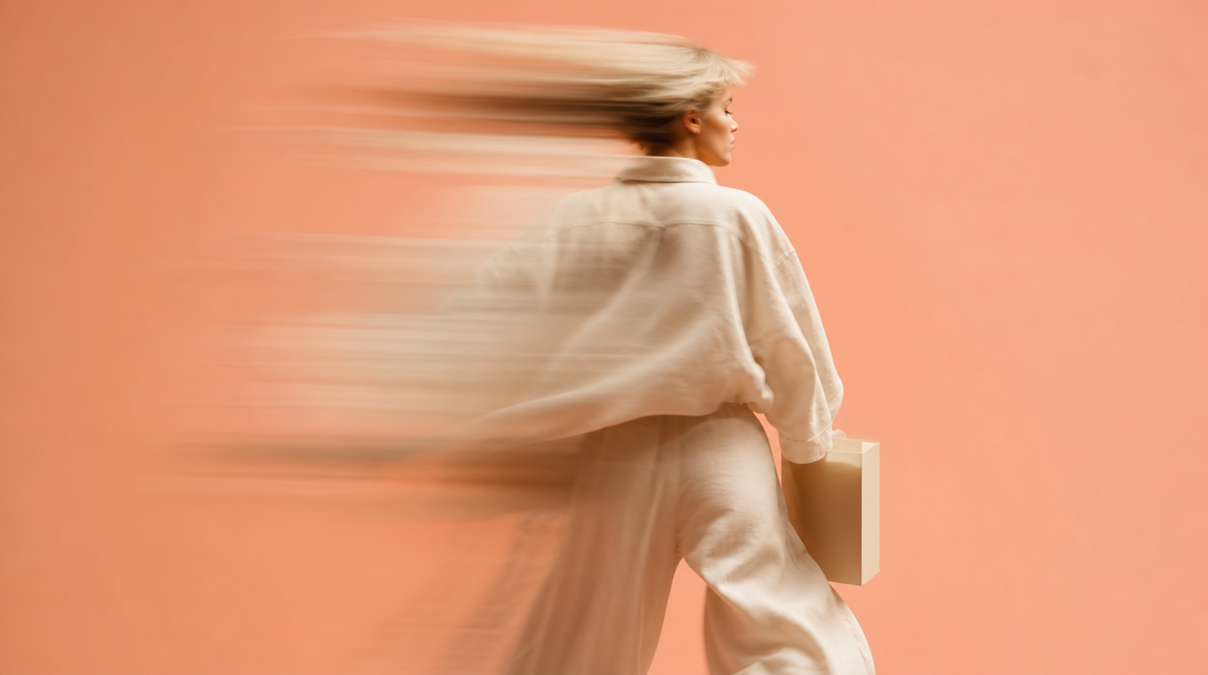 A person mid-spin in a peach-backdrop studio, holding a generic blank-packaged product loosely at waist height, shot from behind at a slight angle,