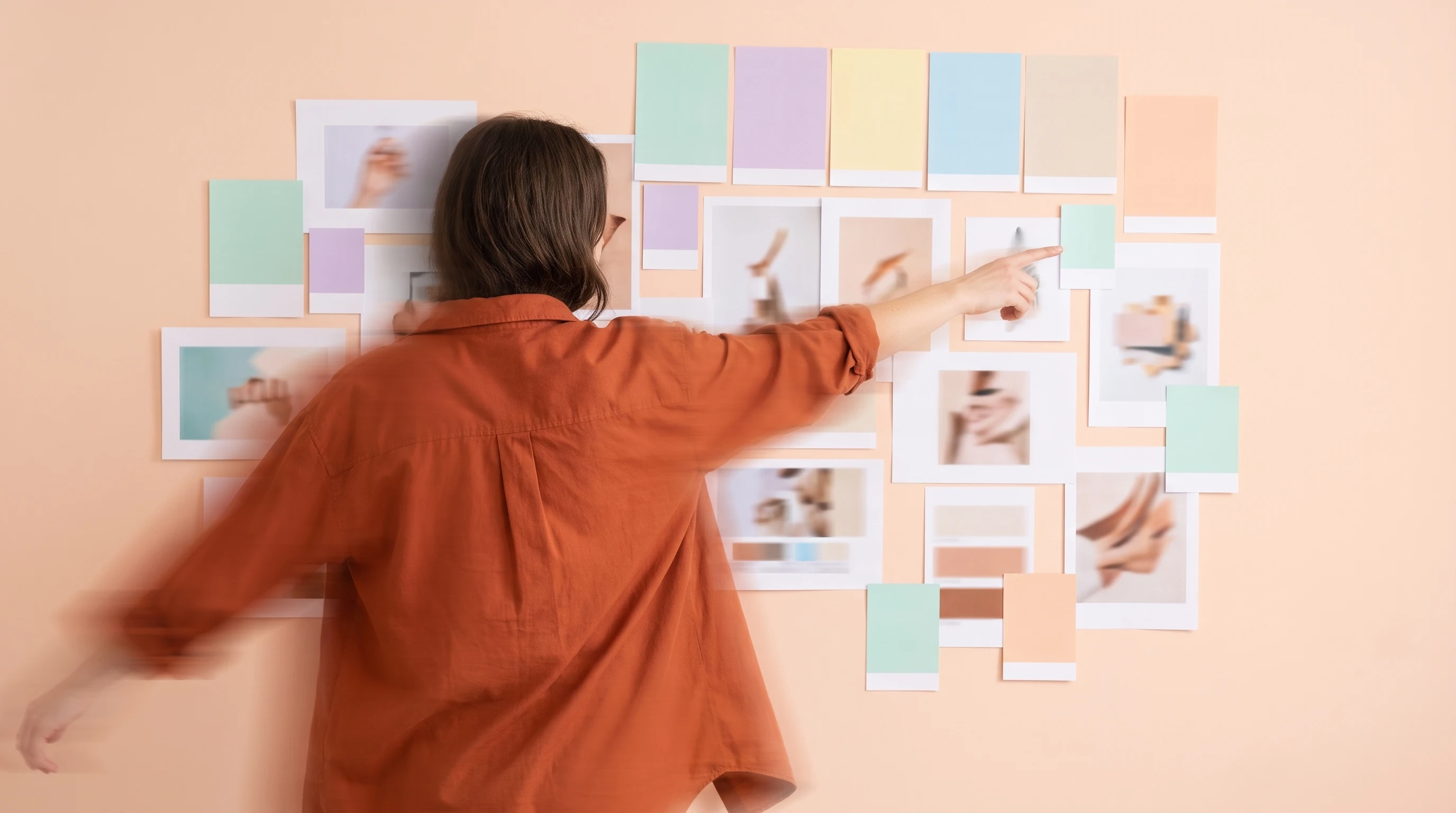 a person mid-turn from behind, arm extended pointing at a wall covered in unbranded campaign printouts and colour swatches, wearing a rust-orange