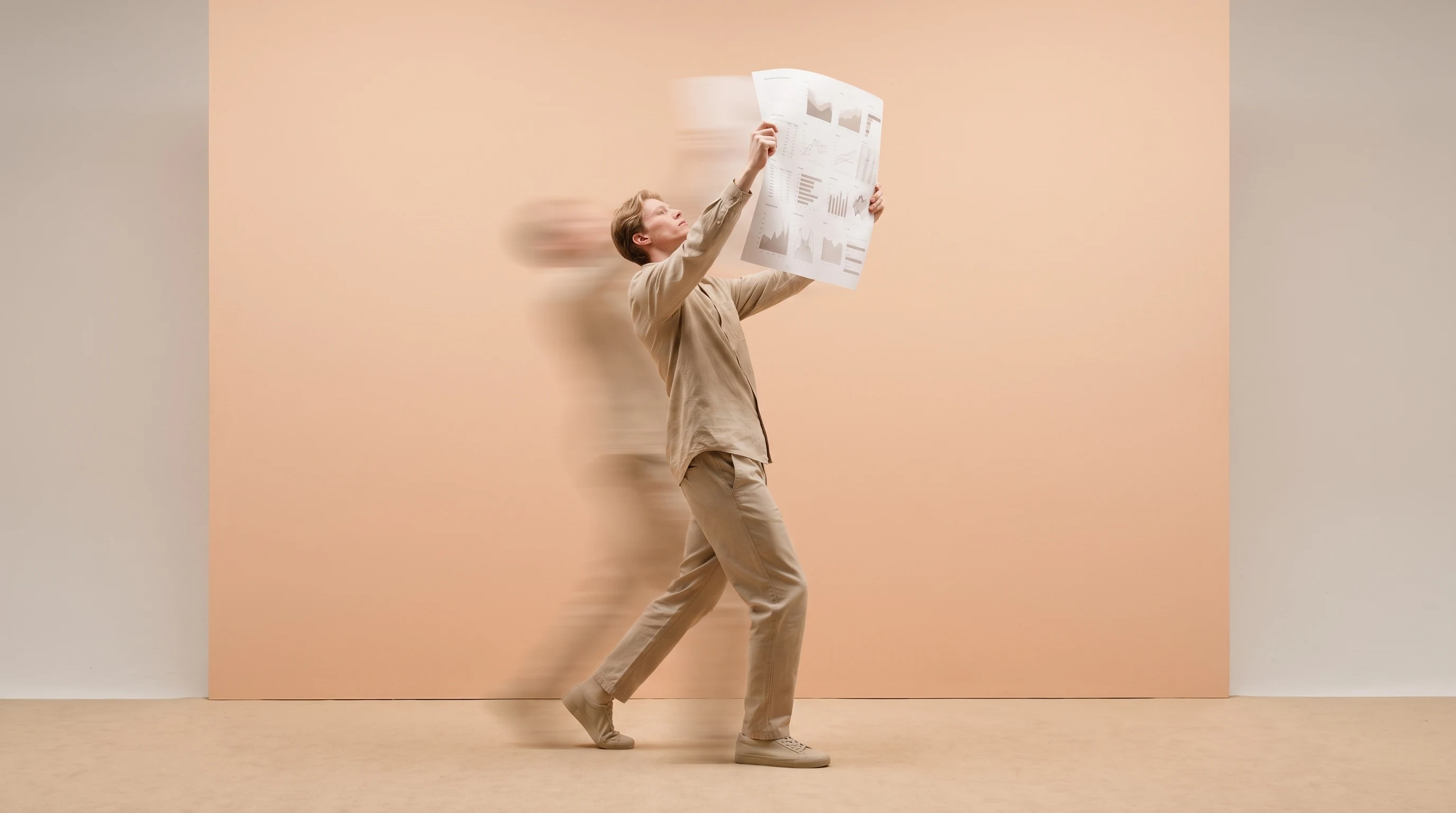 A person mid-step in a sand-toned studio, holding a large unbranded printed chart overhead with both hands, head tilted up reviewing it, shot from