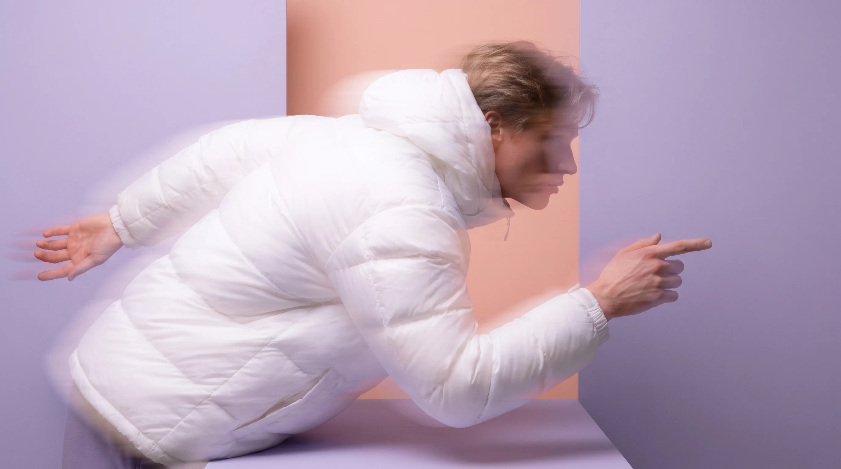 A person in a lilac studio mid-lean over a low unseen surface, one hand pointing forward and one braced behind, framed in tight profile with a peach