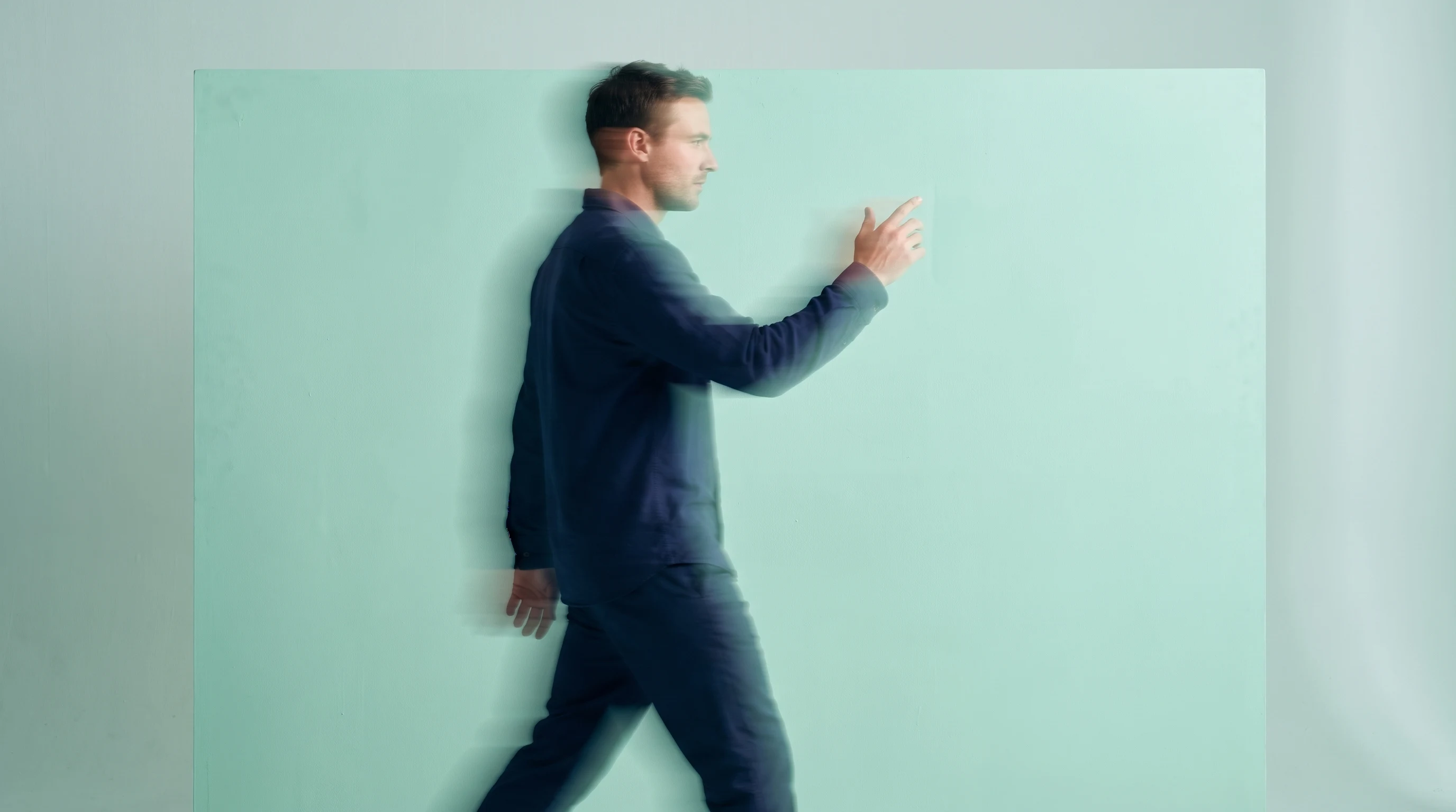 A man mid-step across a mint-backdrop studio in a relaxed navy outfit, profile framing, one hand raised mid-gesture as if presenting something just
