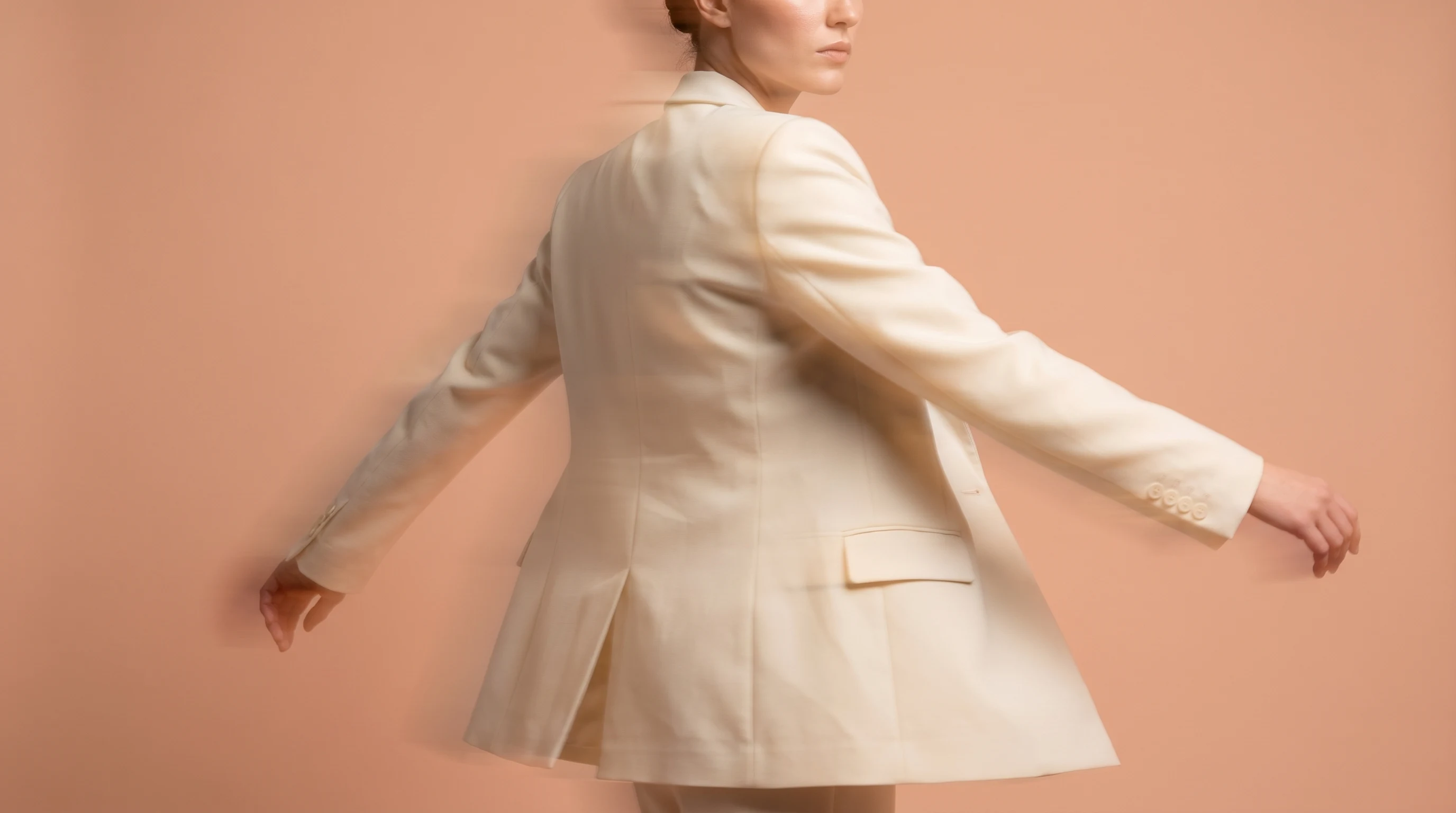 A woman mid-turn in a peach-backdrop studio, arms slightly extended, wearing a tailored cream blazer, three-quarter framing, facing slightly away