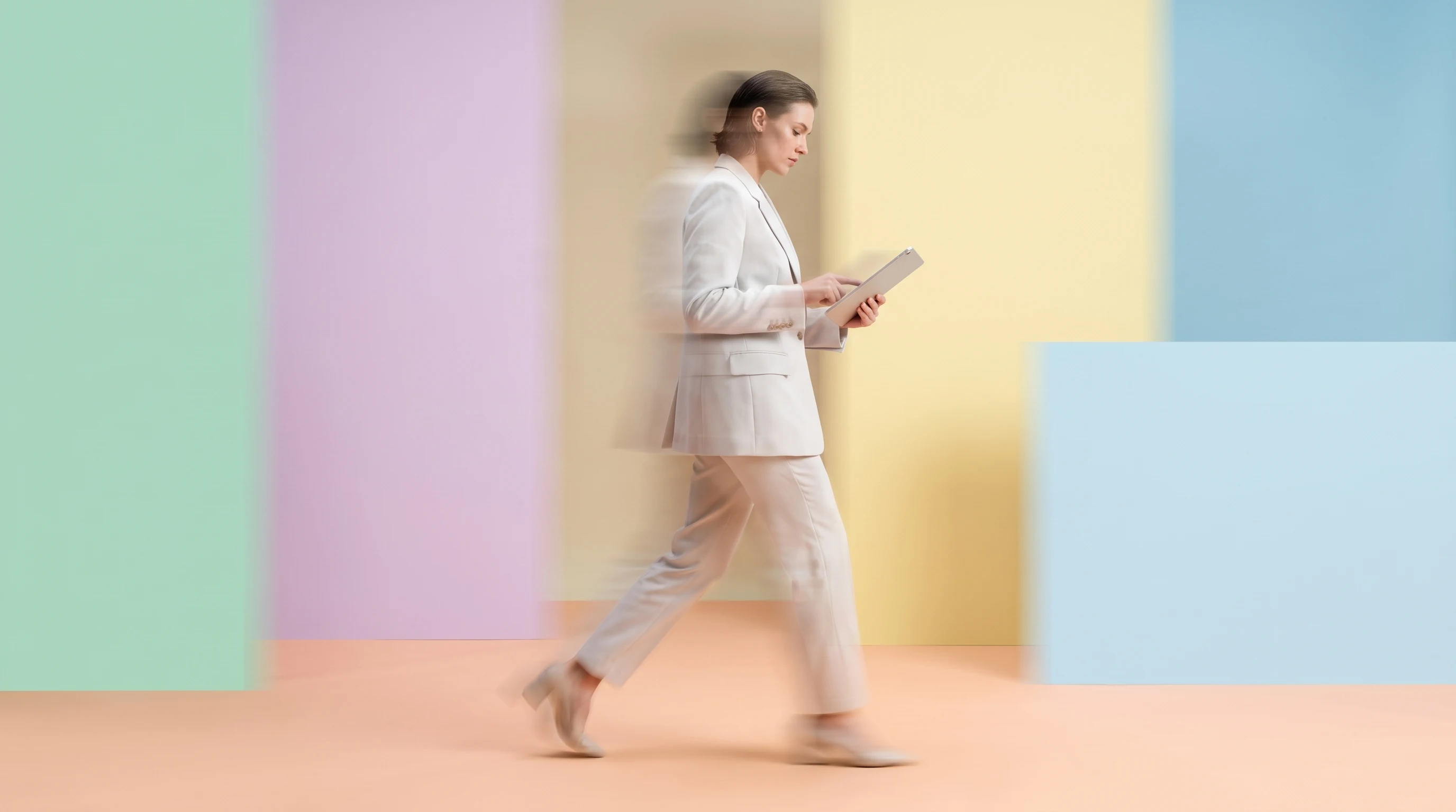 A person mid-step across a peach studio, glancing down at an unbranded tablet held at hip height, profile framing, light outfit with a structured