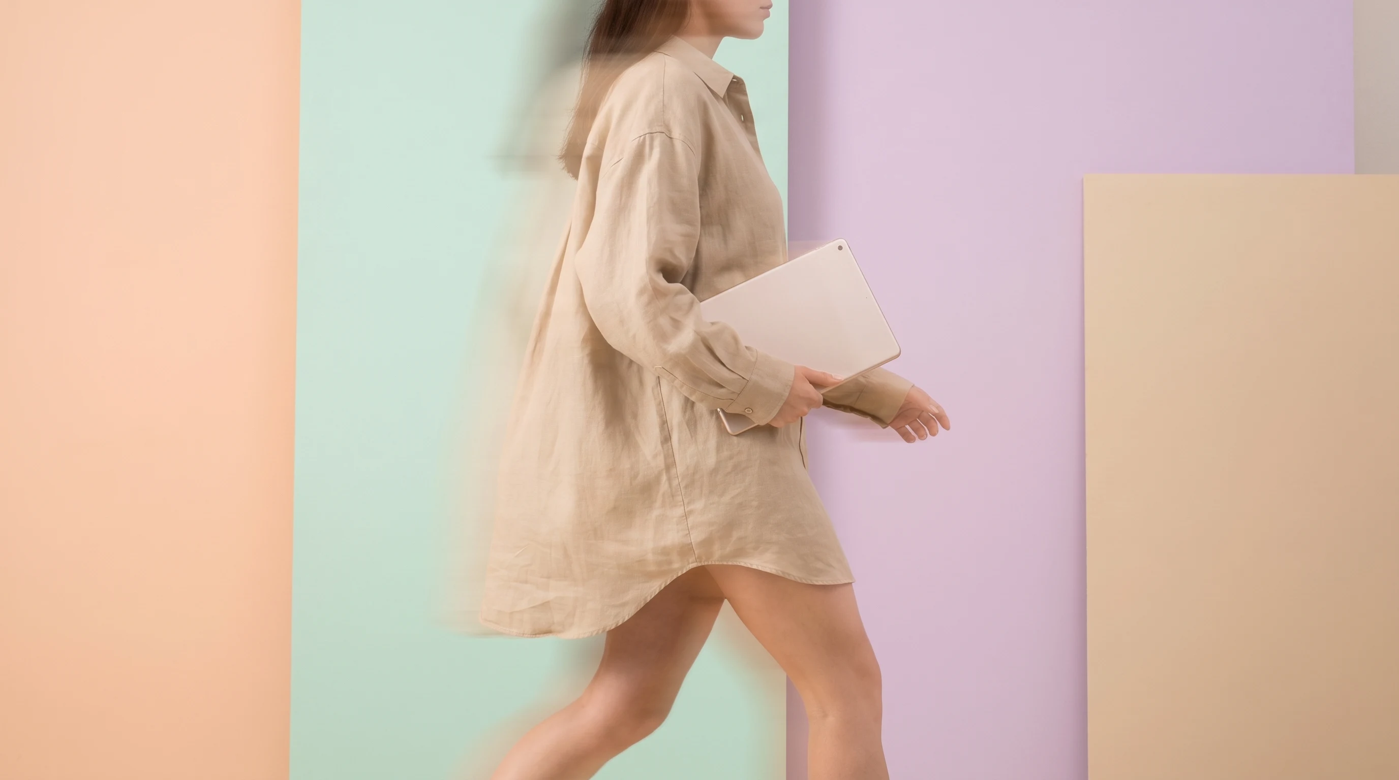 a person mid-step in a peach studio, holding a blank unbranded tablet face-out toward camera, wearing an oversized sand linen shirt, framed from