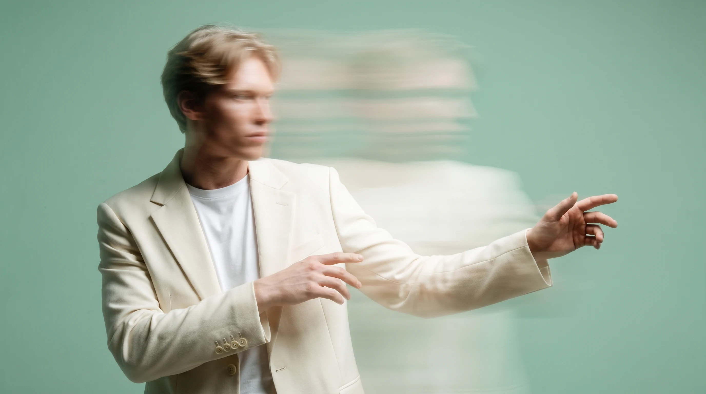 a person mid-turn in a mint studio, gesturing toward an off-frame screen, wearing a structured cream blazer over a white tee, three-quarter framing