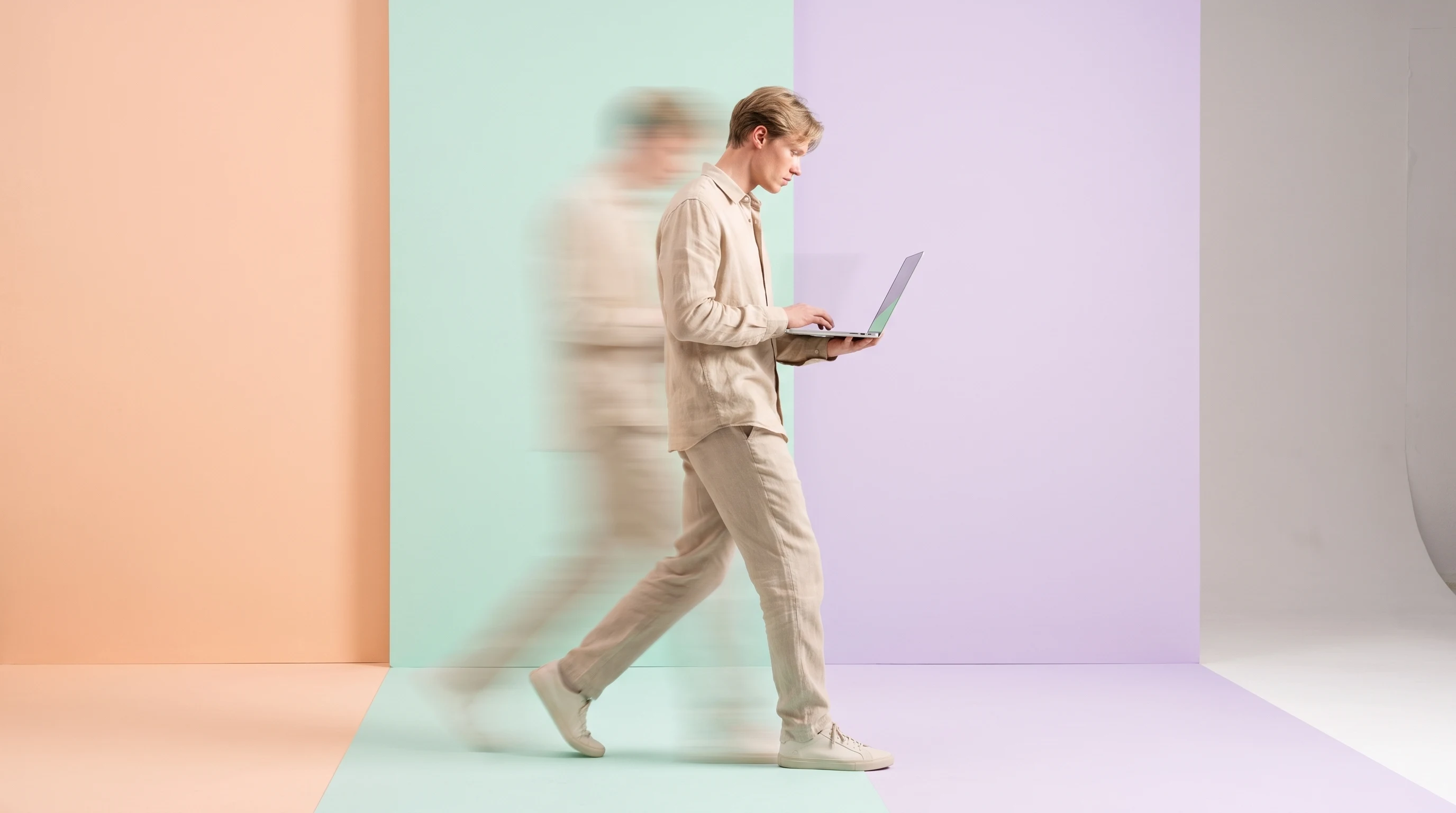 A person mid-step across a peach-backdrop studio holding a slim open laptop at hip height, glancing down at the screen, dressed in a relaxed sand