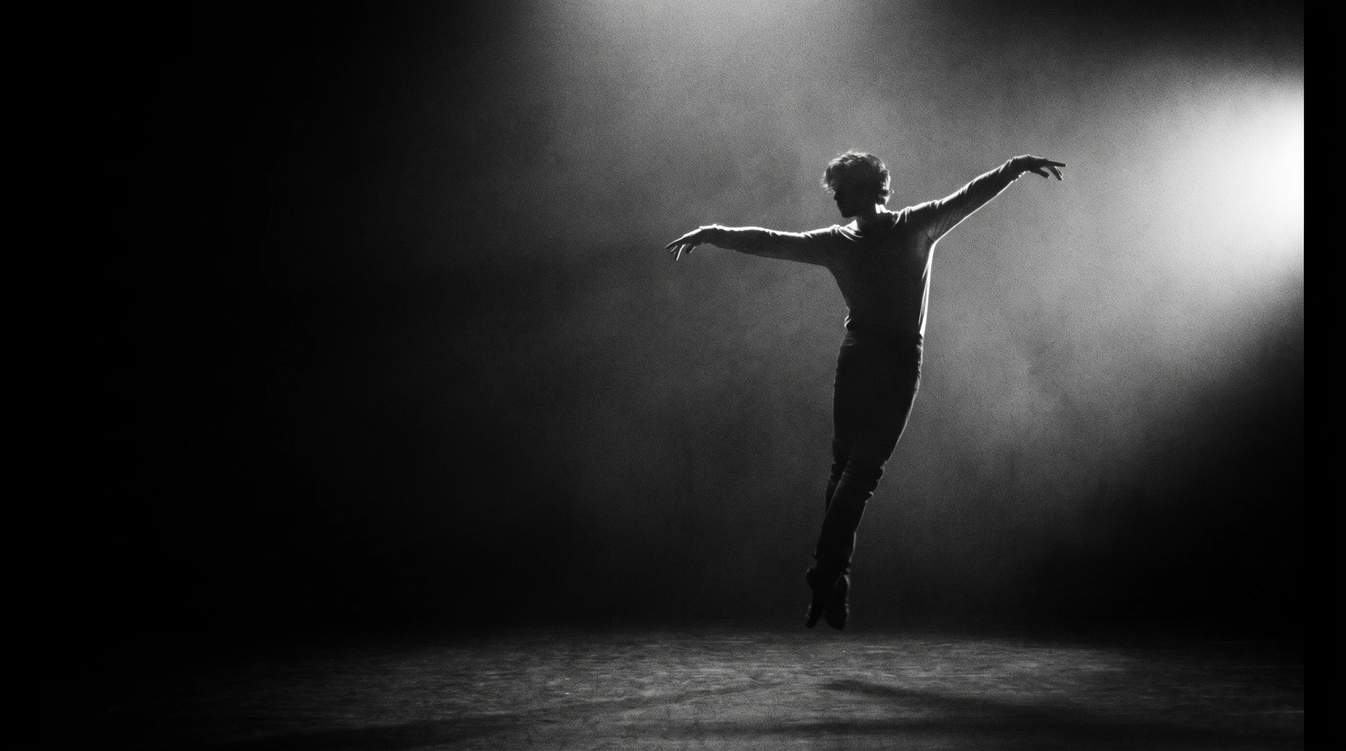 A performer mid-leap in a dark sparse studio, arms wide, backlit by a single off-frame light source as if caught mid-take for a video